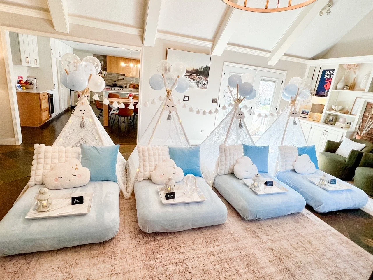 Baby sleepover setup with four beds on a pink rug, decorated with cloud pillows, light blue cushions, and teepees with balloons and cute hanging plush toys, in a cozy living room.