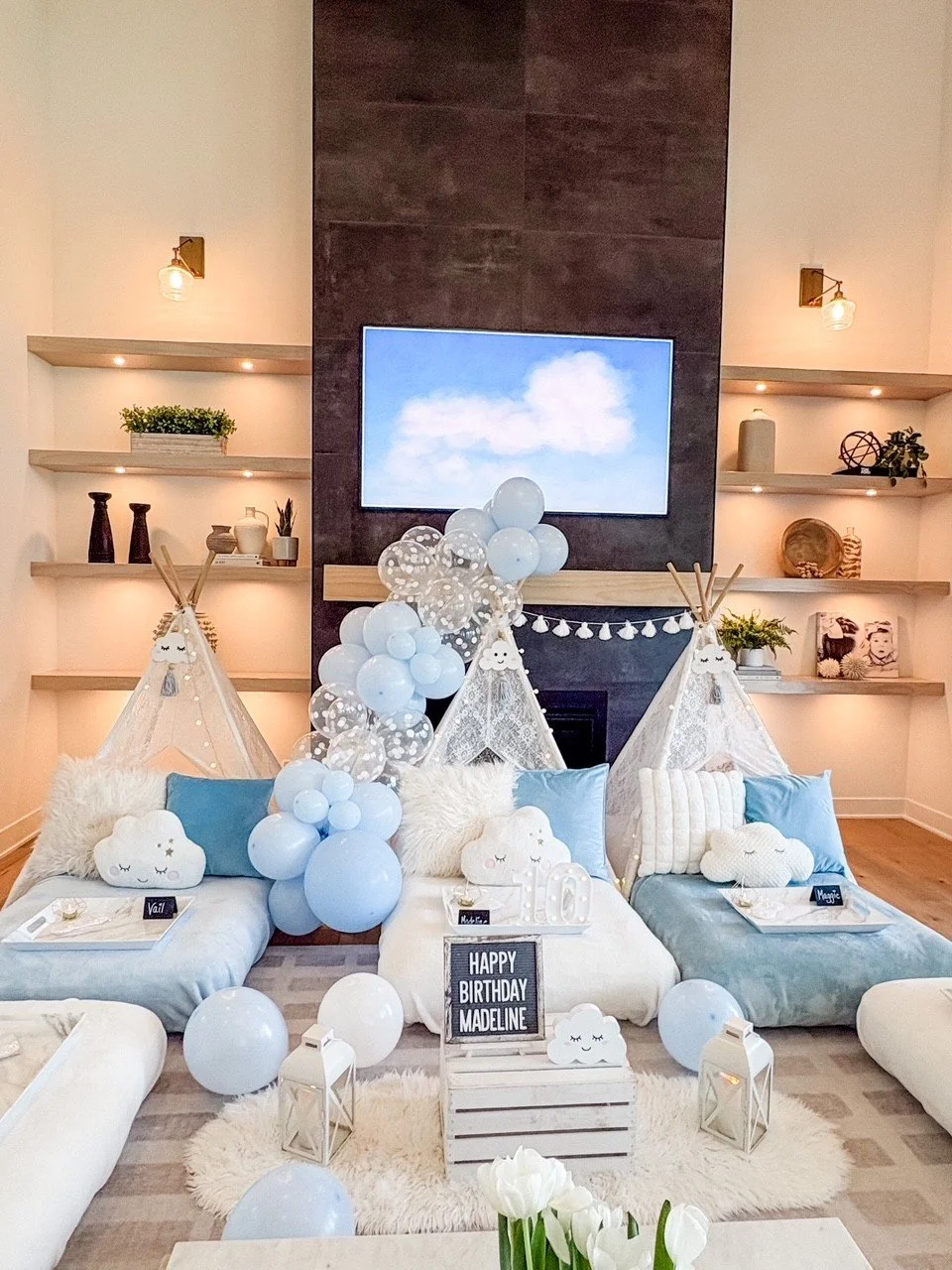 Decorated living room with white and blue themed birthday setup featuring teepees, balloons, cloud pillows, lanterns, and a sign reading 'Happy Birthday Madeline'.
