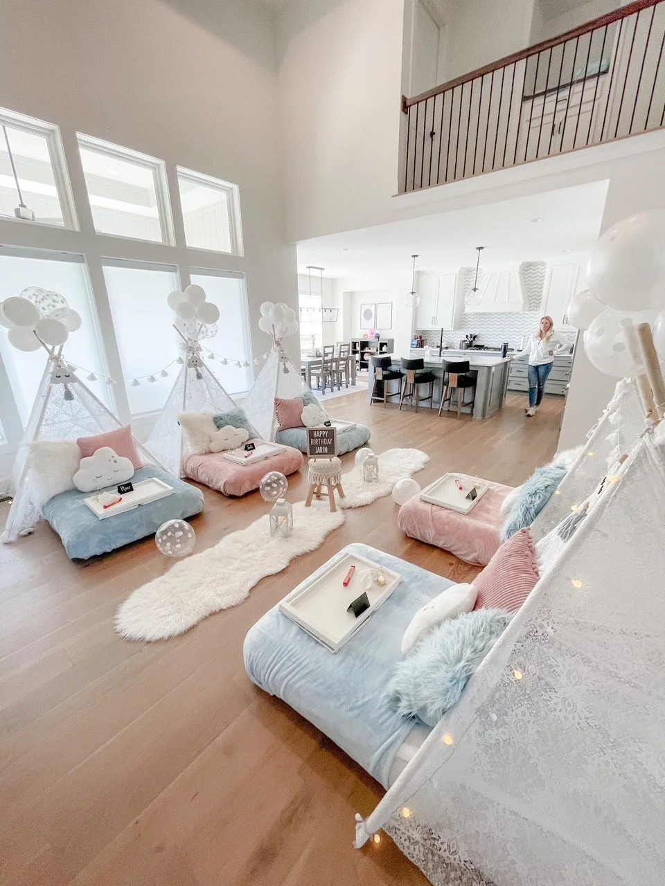 Indoor birthday party setup with pastel-colored couches and teepees decorated with balloons and fairy lights, located in a bright open living space.