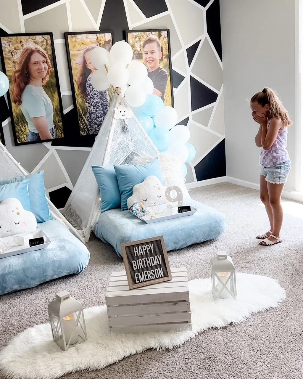 A girl in pajamas appears surprised or excited at a decorated birthday celebration for Emerson, with pillows, plush cloud decor, balloons, framed photos, and a sign that reads 'Happy Birthday Emerson'.