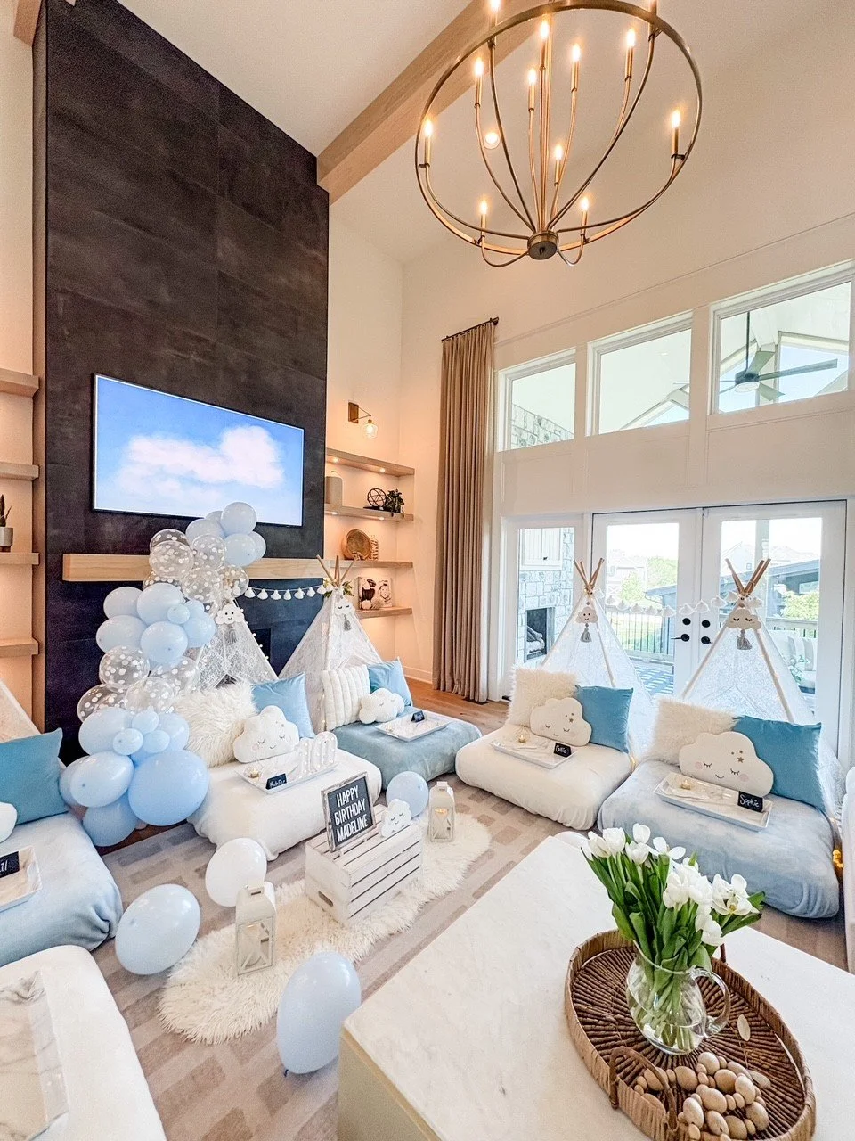 Living room decorated for a birthday party with cloud and teepee themed decorations, balloons, plush pillows, and a vase of white tulips.