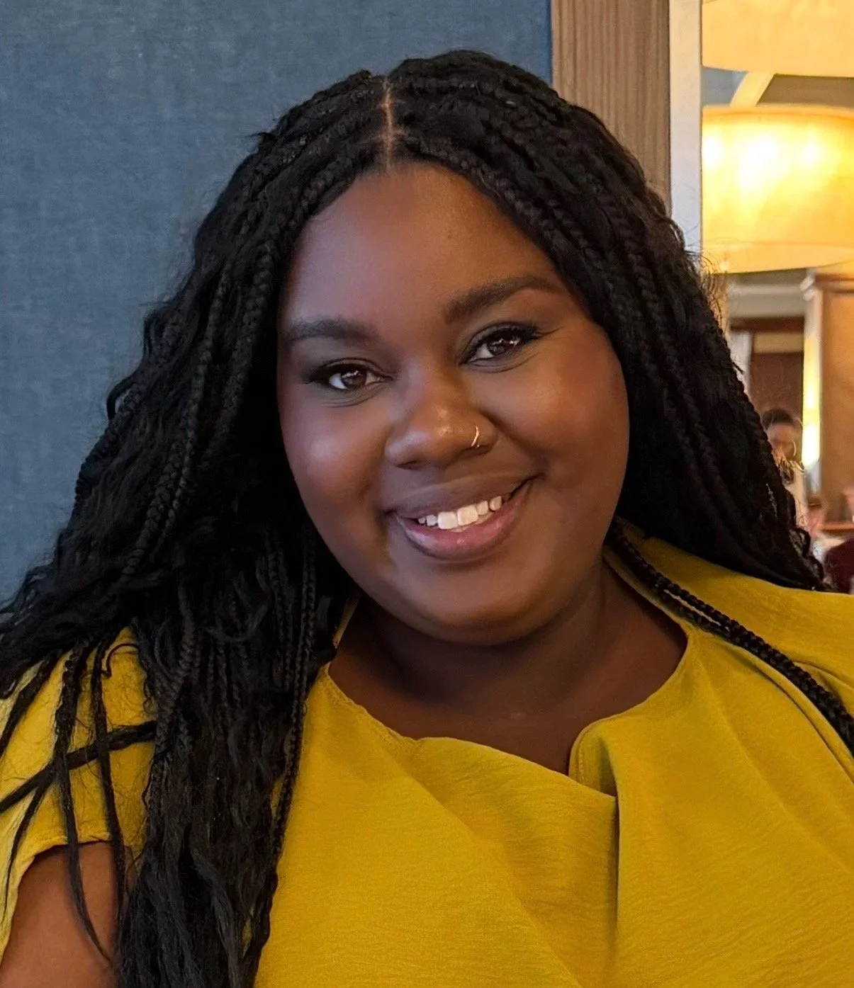 A woman with dark hair styled in braids, wearing a nose ring and yellow top, looking at the camera with a slight smile.