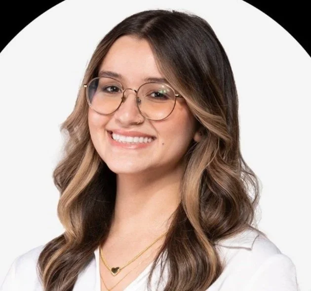 A young woman with long, wavy brown hair, wearing glasses with a thin metal frame, a white top, and layered gold necklaces, smiling in front of a white background.