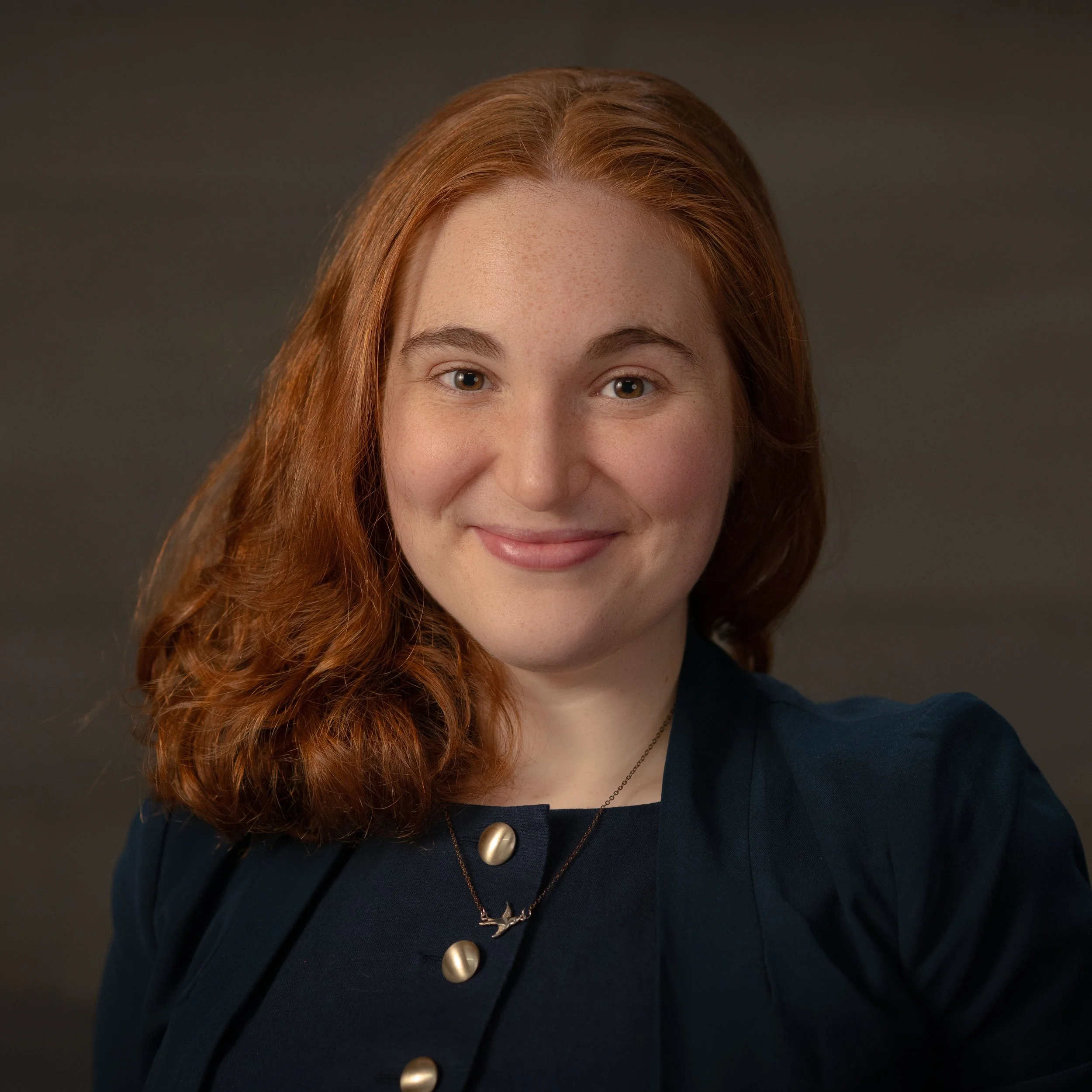 A young woman with curly red hair smiling at the camera, wearing a black blazer and a patterned blouse, against a dark background.