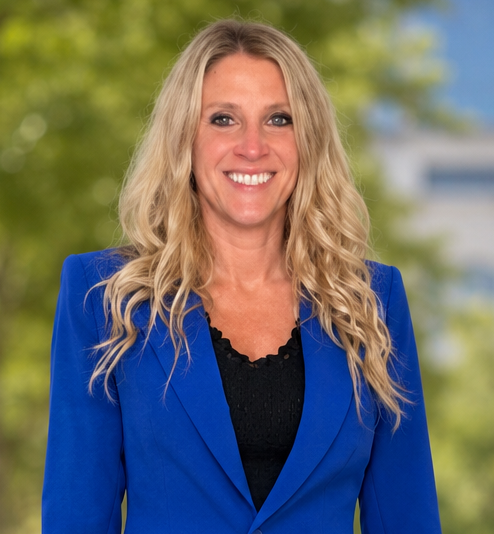 A woman with long wavy blonde hair smiling outdoors, wearing a black leather jacket, with a body of water and a hill in the background.
