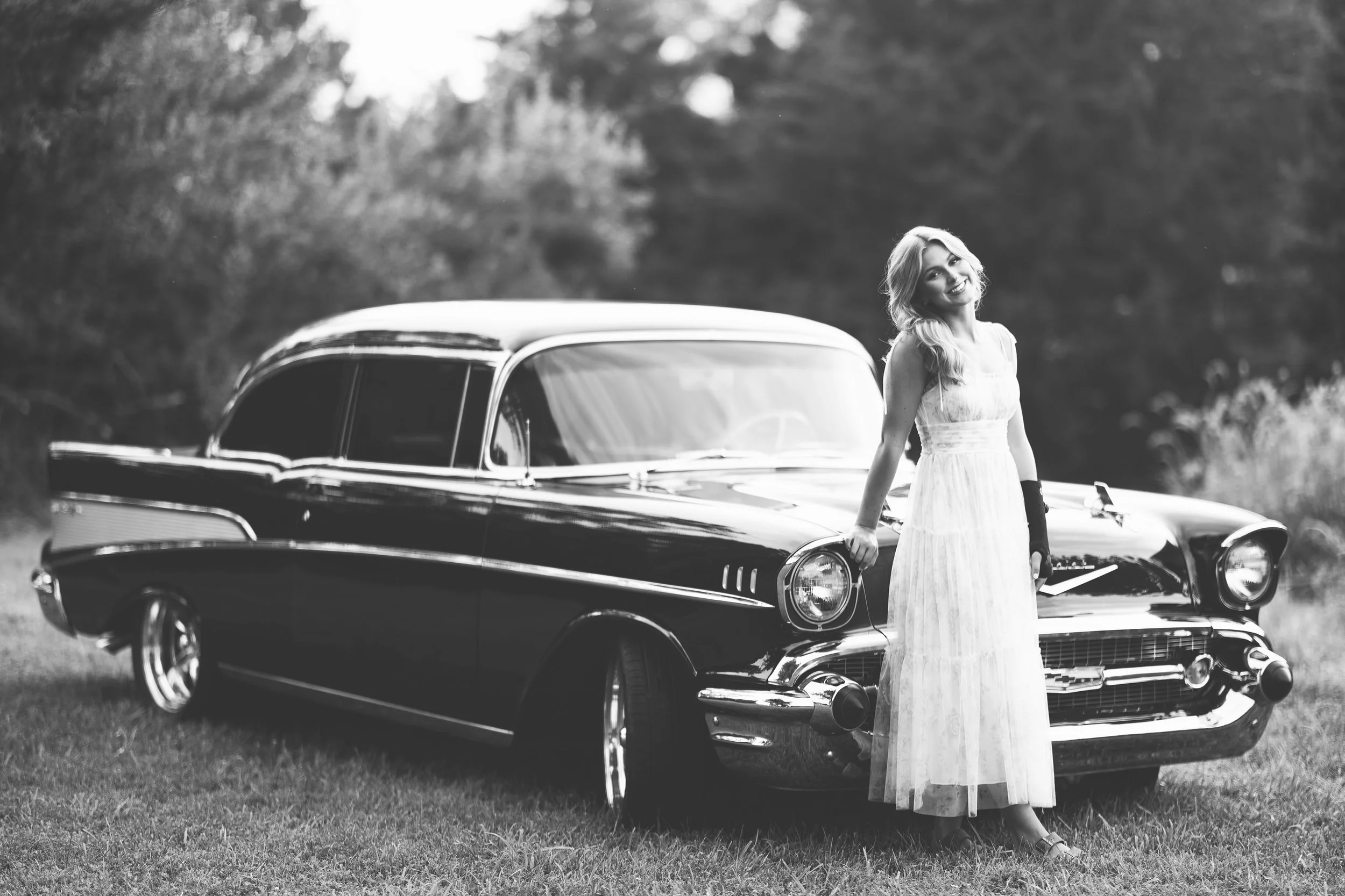 High school senior girl in a long white dress posing beside a classic Chevrolet car in a black-and-white portrait.