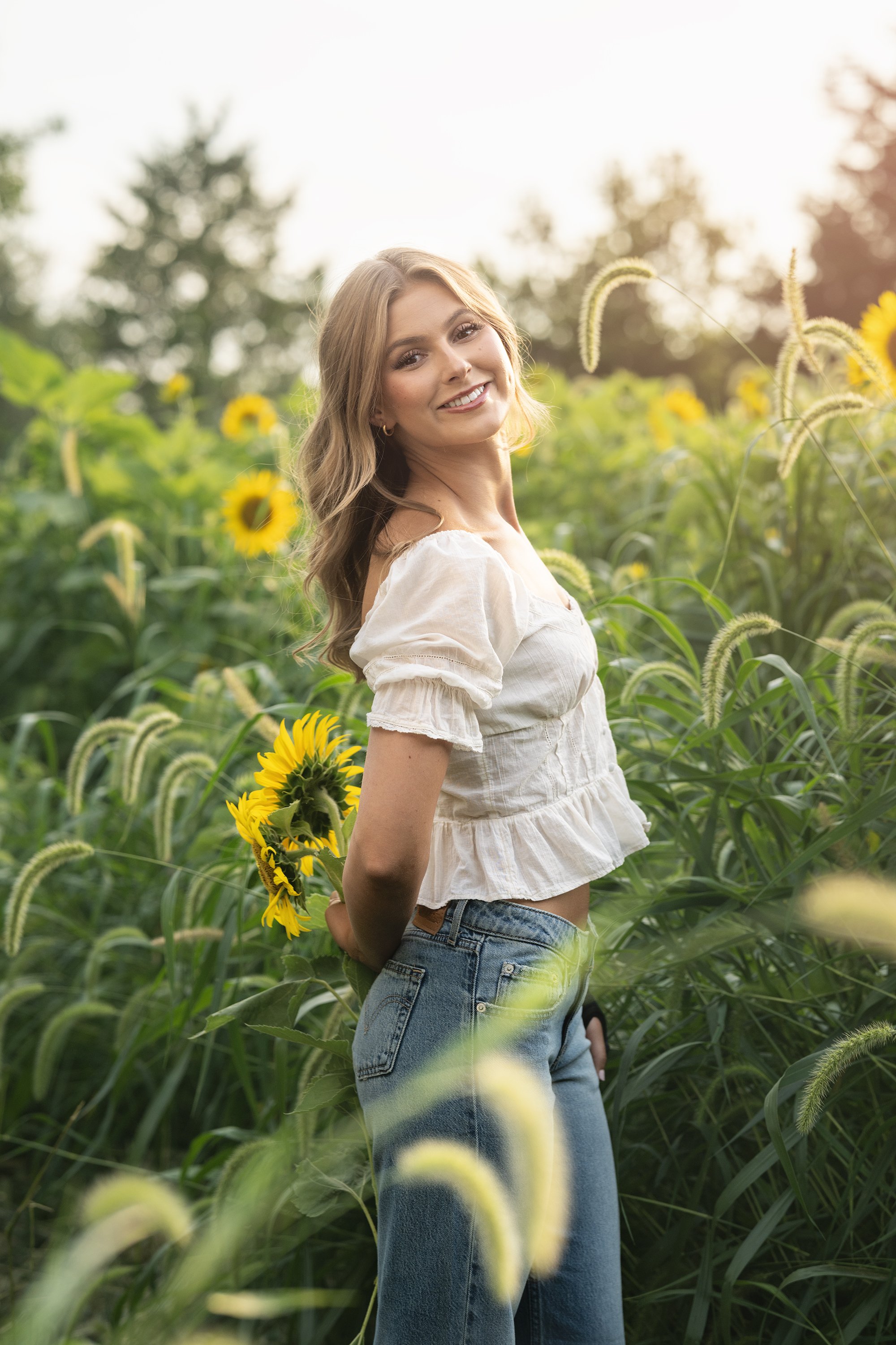 Class of 2027 Senior Girl blue jeans cream top in field of sunflowers at goldenHour in Marion Illinois