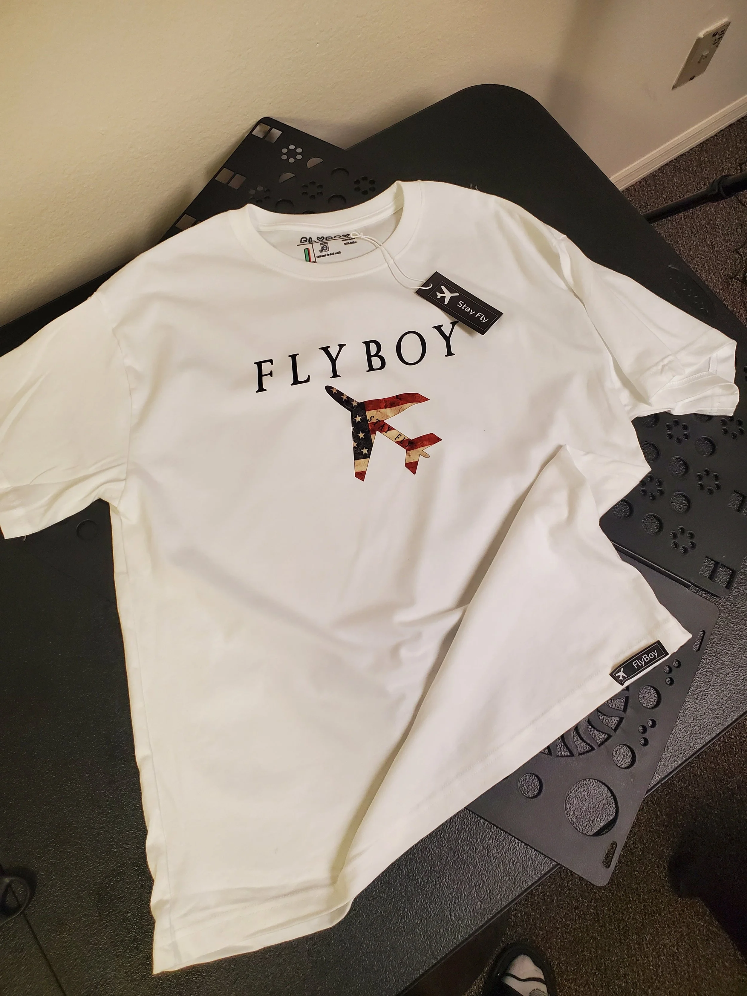 A white T-shirt with the word 'FLYBOY' and a stylized airplane logo, which features an American flag design, on it. The T-shirt is laying on a black table, with a tag attached that reads 'Stay Fly' and includes a small airplane icon.