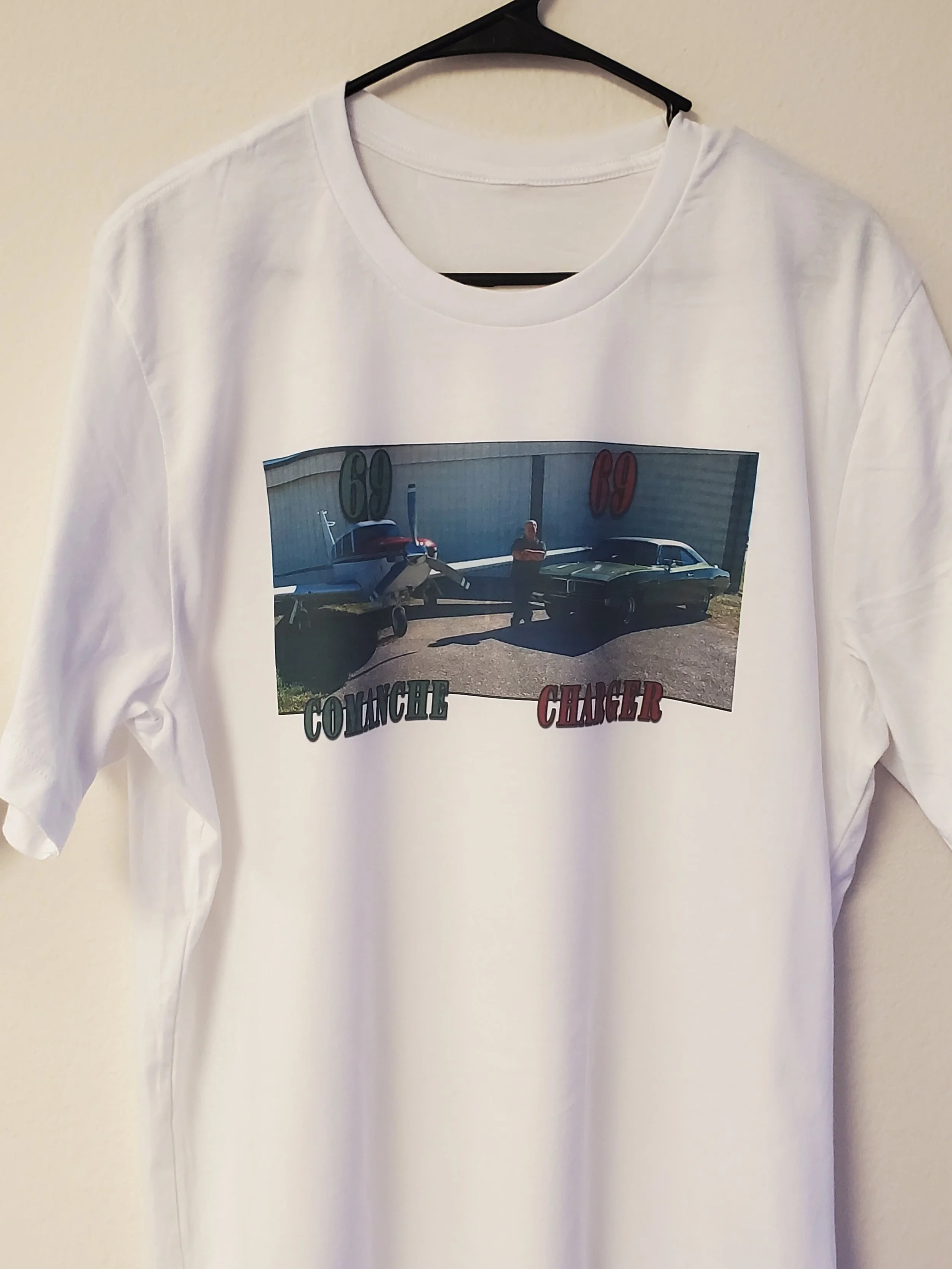 White T-shirt featuring a photo of a man standing between a vintage muscle car and a vintage jet aircraft in front of a hangar, with the numbers '60' and '69' above, and the words 'Collision' and 'Charger' underneath.