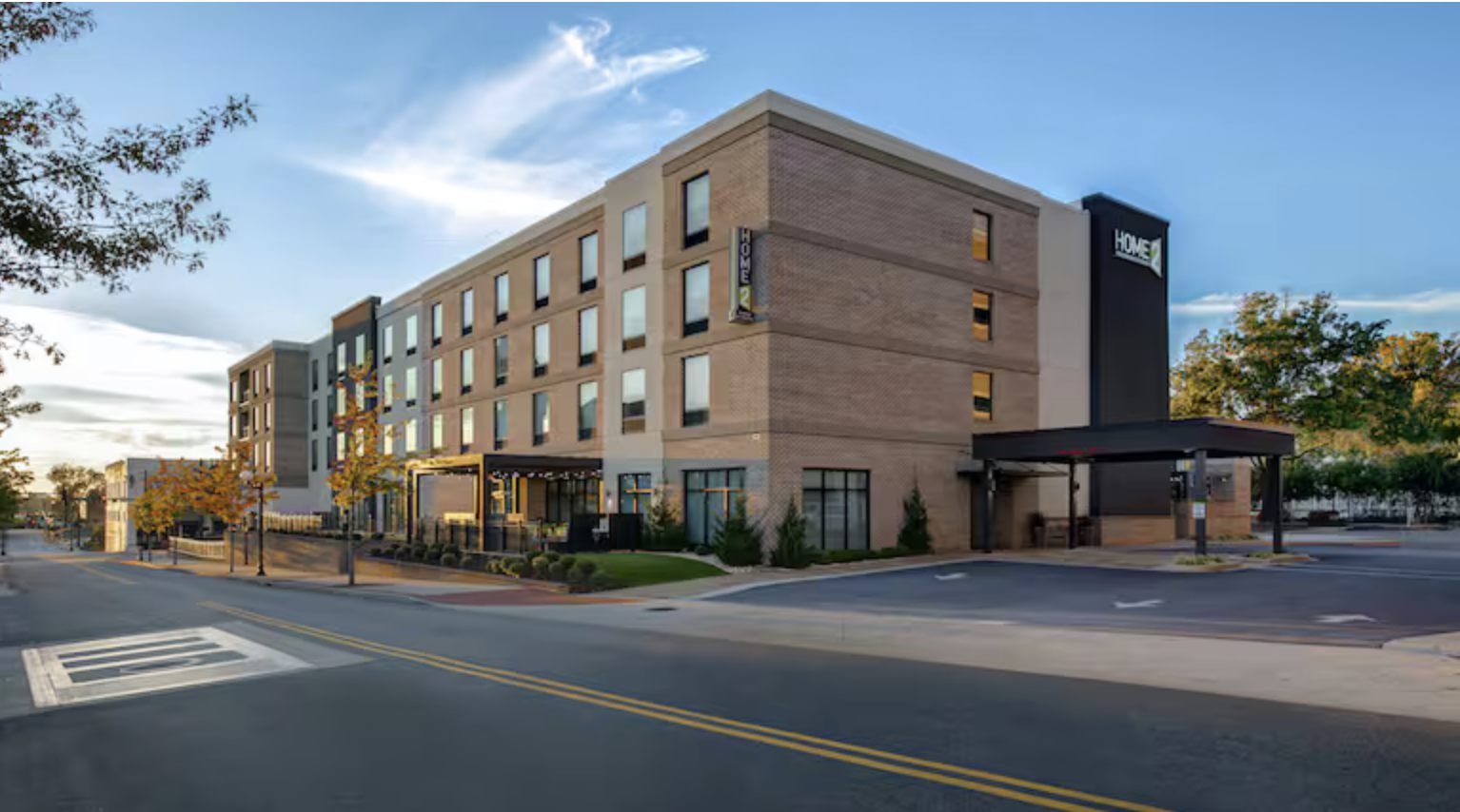 Modern brick hotel building with large windows and a covered entrance, surrounded by trees and a quiet street, under a blue sky.