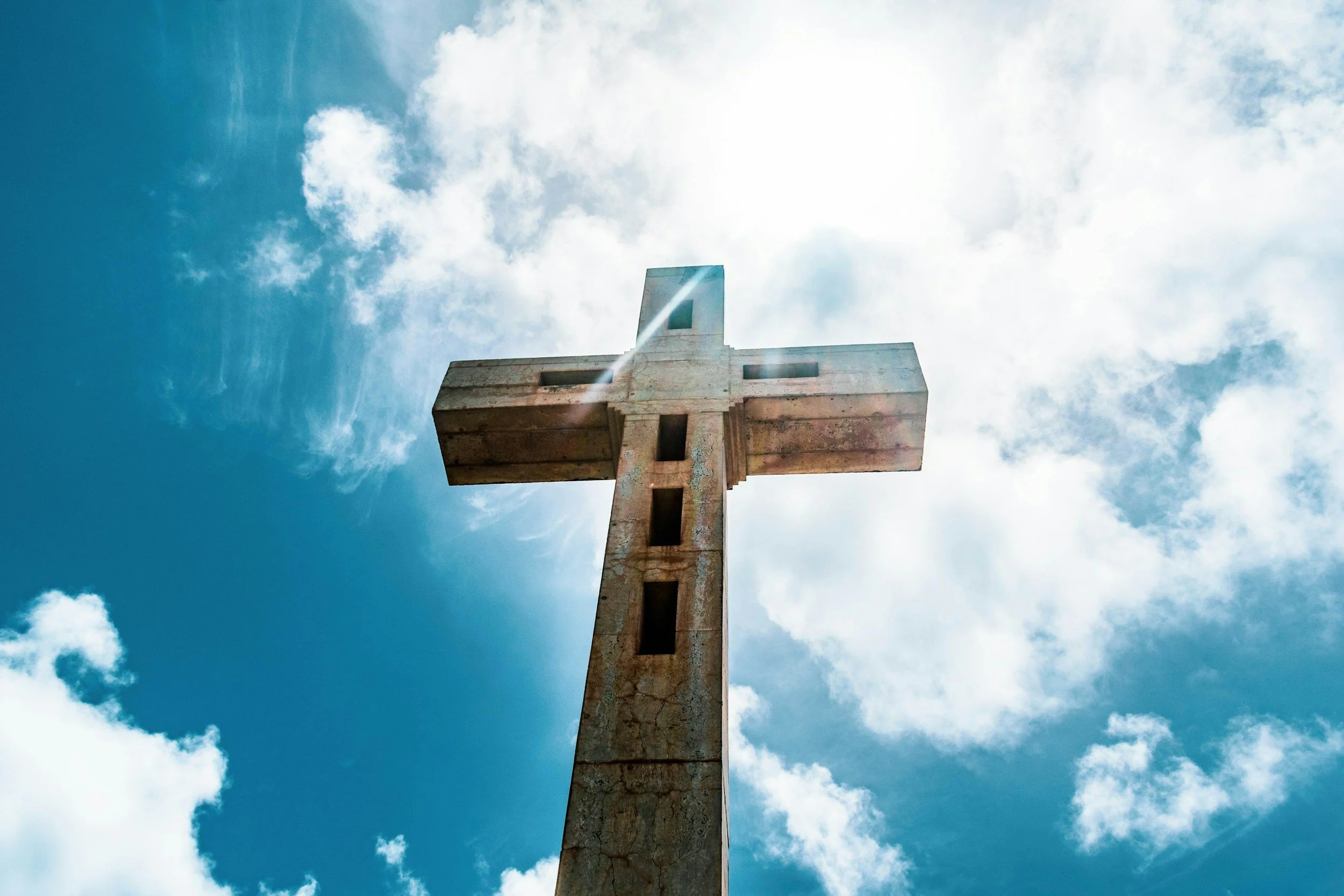 A large stone cross monument against a partly cloudy sky with sunlight shining through.