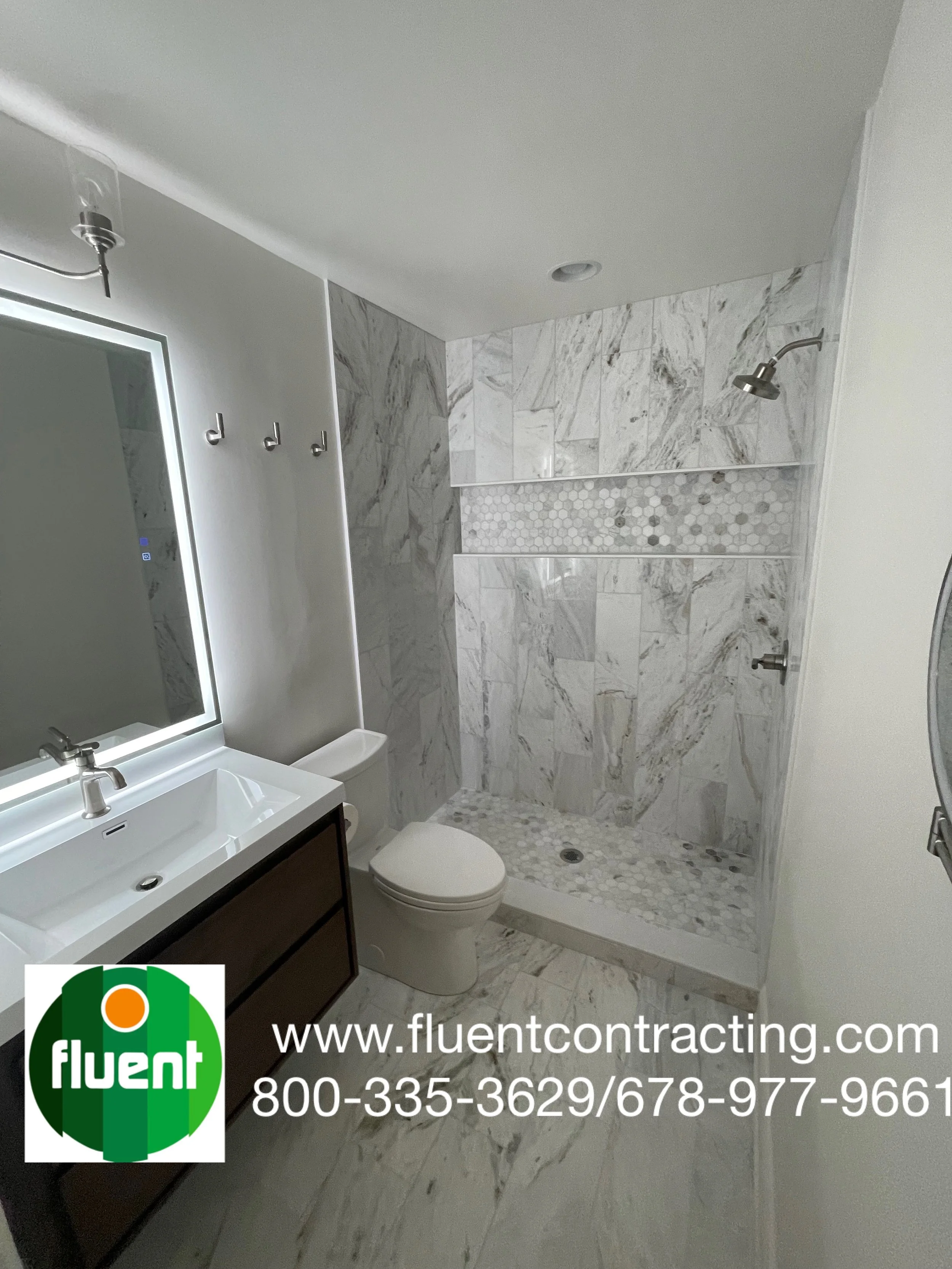 Modern bathroom with marble tiles, glass shower enclosure, white sink, and a mirror with LED lighting, featuring a toilet and decorative wall hooks.