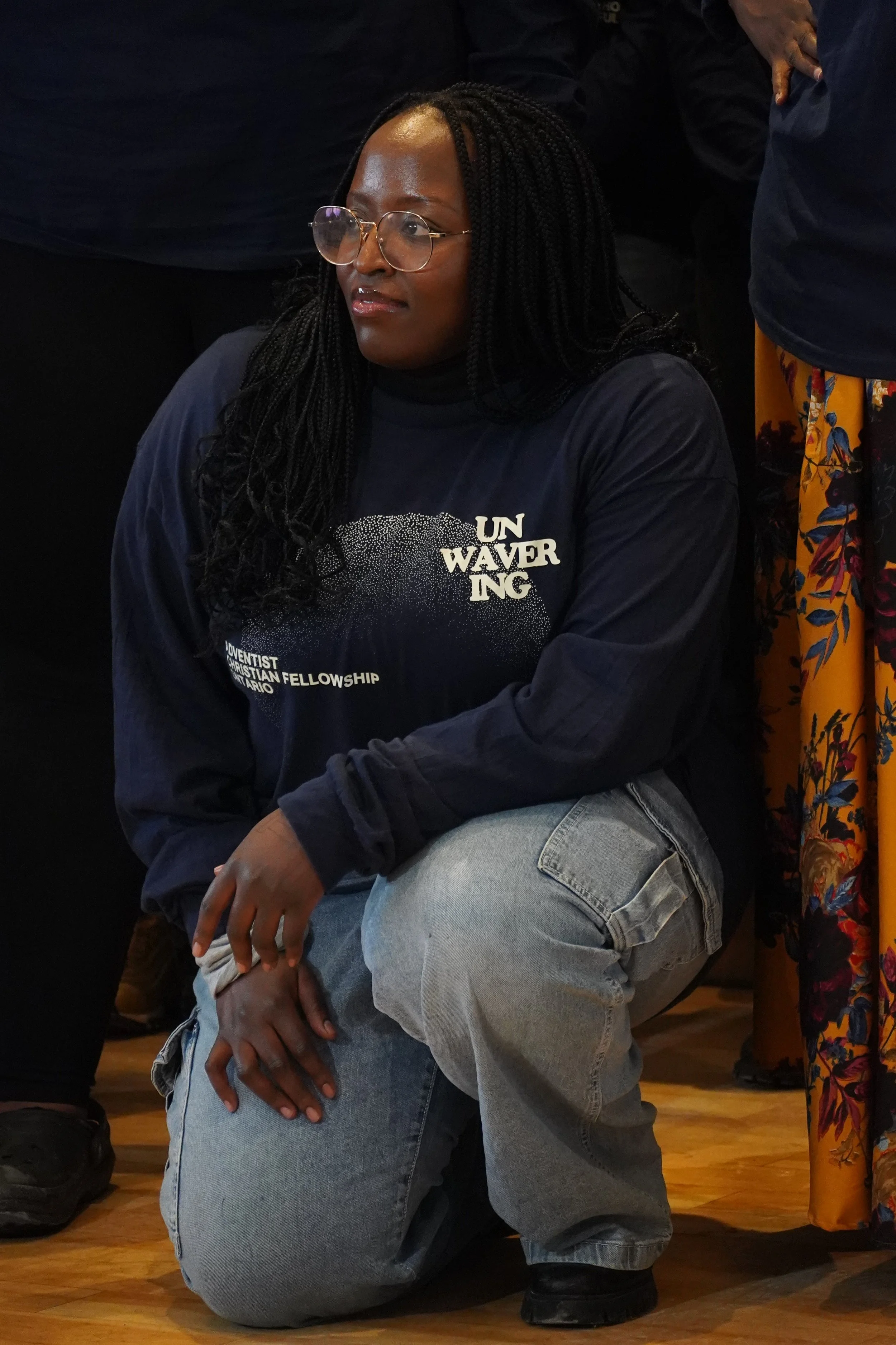 A woman with glasses, long black braids, wearing a navy sweatshirt and light jeans, kneeling on a wooden floor in a group setting.