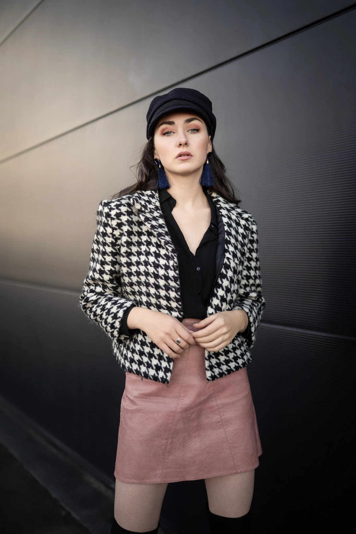 Jeune femme élégante portant un blazer à motif pied-de-poule, un chapeau noir, une jupe en daim rose, et des boucles d'oreilles bleues, posant contre un mur sombre. style urbain, mode chic.