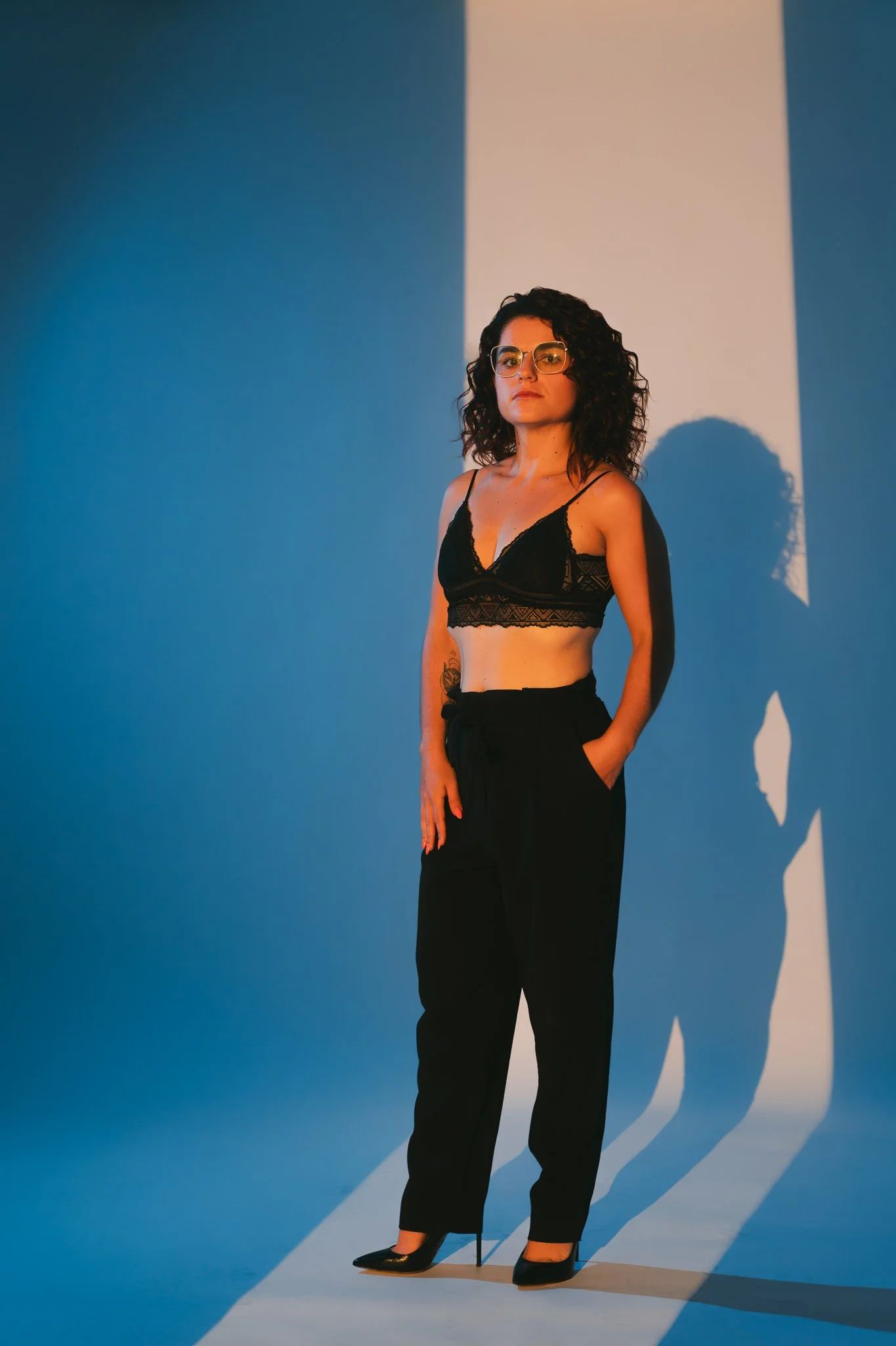 Une femme portant des lunettes, un haut noir en dentelle et un pantalon noir, se tient dans un studio avec un fond bleu et blanche, créant des ombres nettes de sa silhouette.