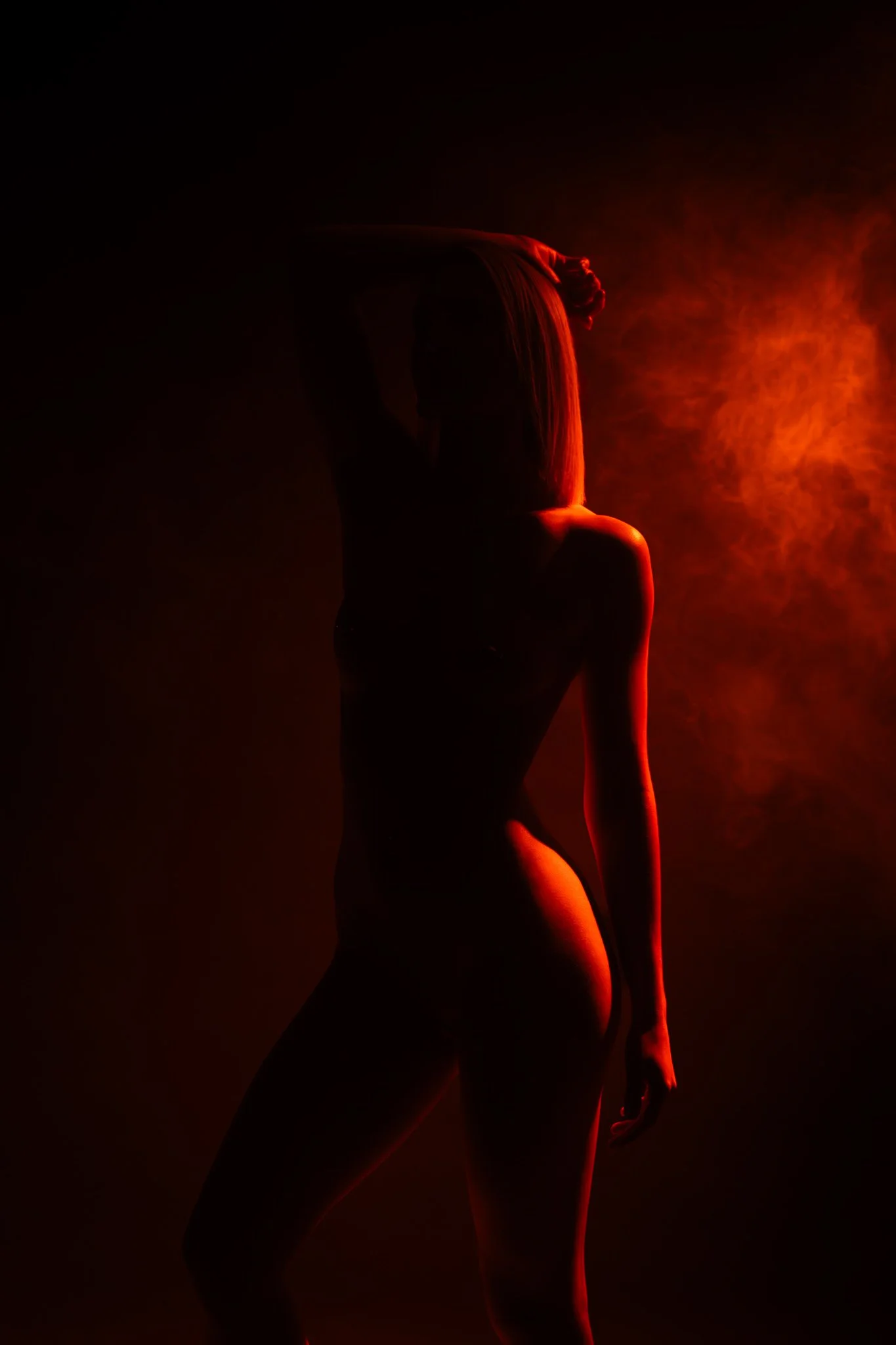 Silhouette d'une femme nue, éclairée par une lumière rouge-orangé, avec de la fumée ou de la vapeur en arrière-plan, créant une atmosphère mystérieuse et artistique.