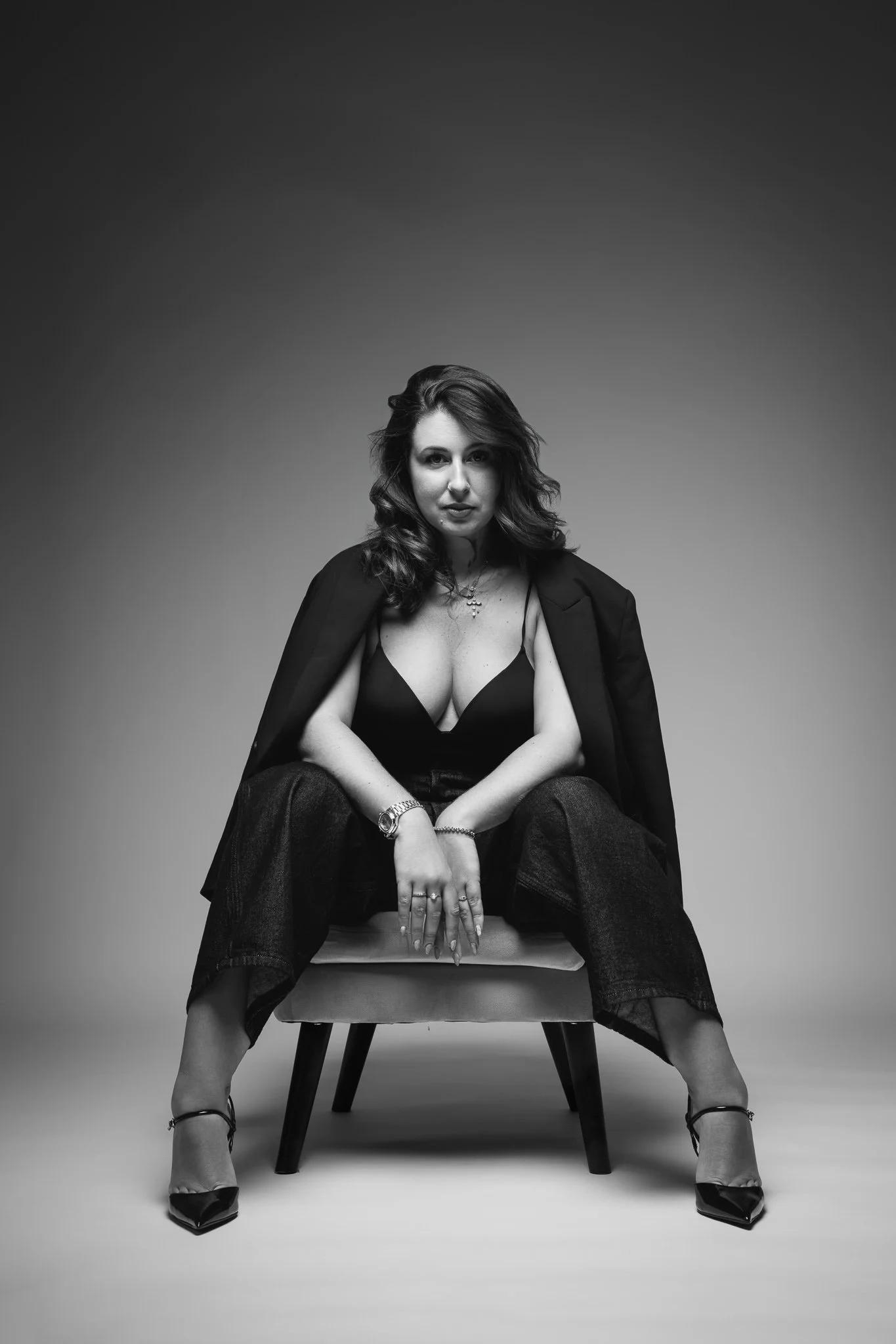 Une femme assise sur une chaise en cuir, portant un blazer noir, une robe noire, des talons hauts et des bijoux, avec une expression confiante, dans une photo en noir et blanc.