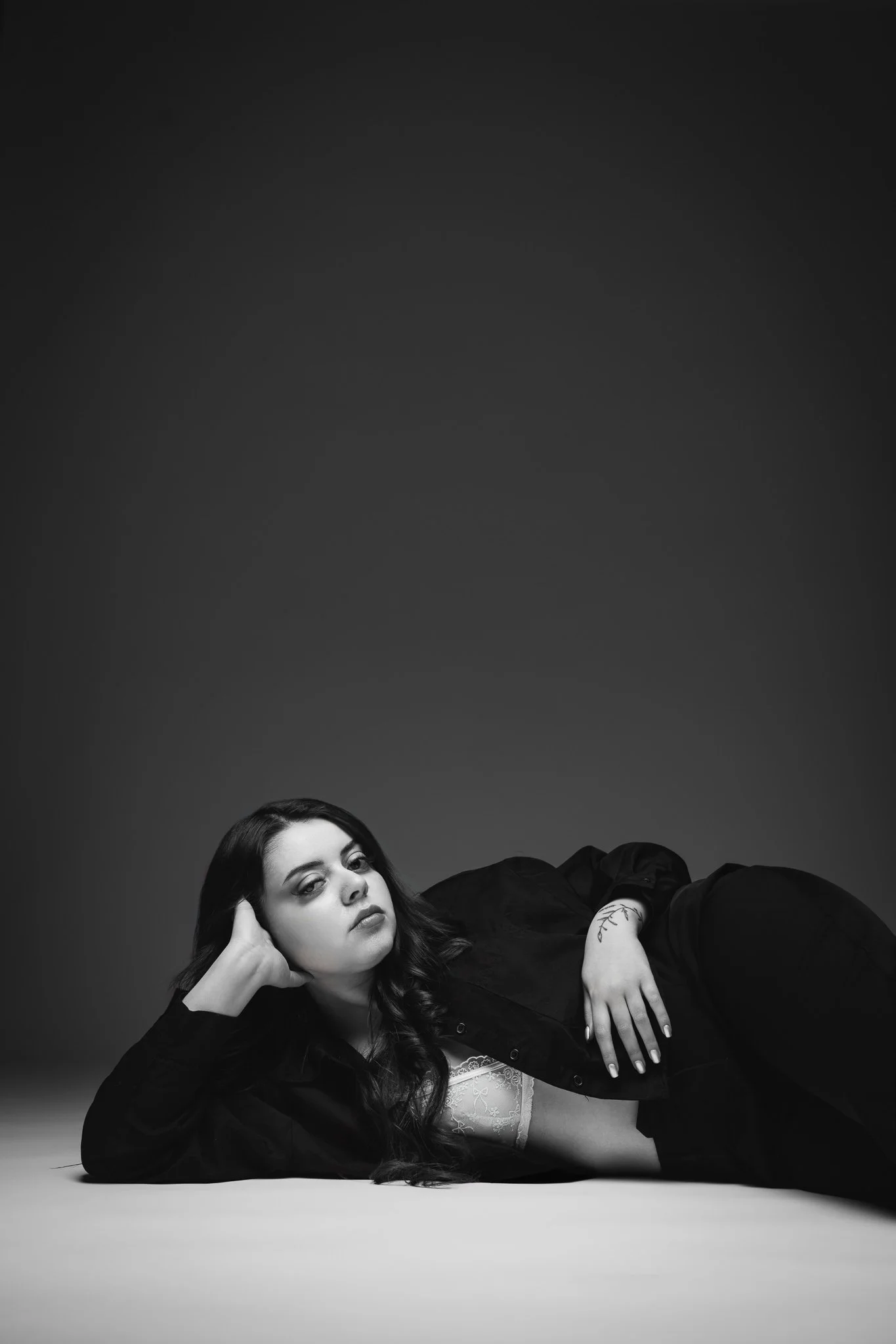 Une femme aux cheveux bouclés, assise au sol, repose sa tête sur sa main gauche, portant une veste noire et un corsage blanc avec des motifs floraux, dans une ambiance monochrome