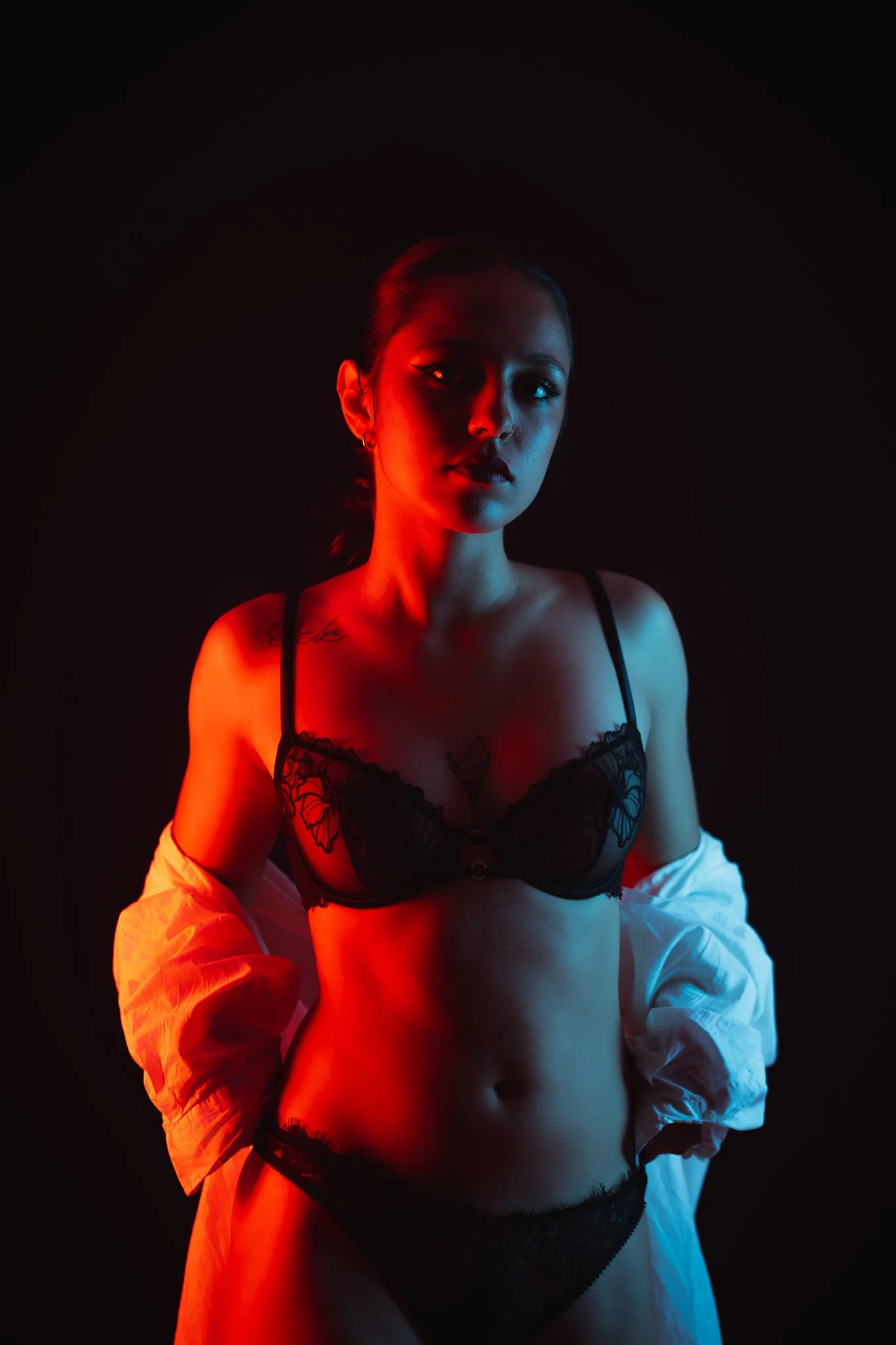 Jeune femme portant des vêtements en lingerie noire et une chemise blanche en arrière-plan, éclairée par des lumières rouges et bleues, contre un fond noir.