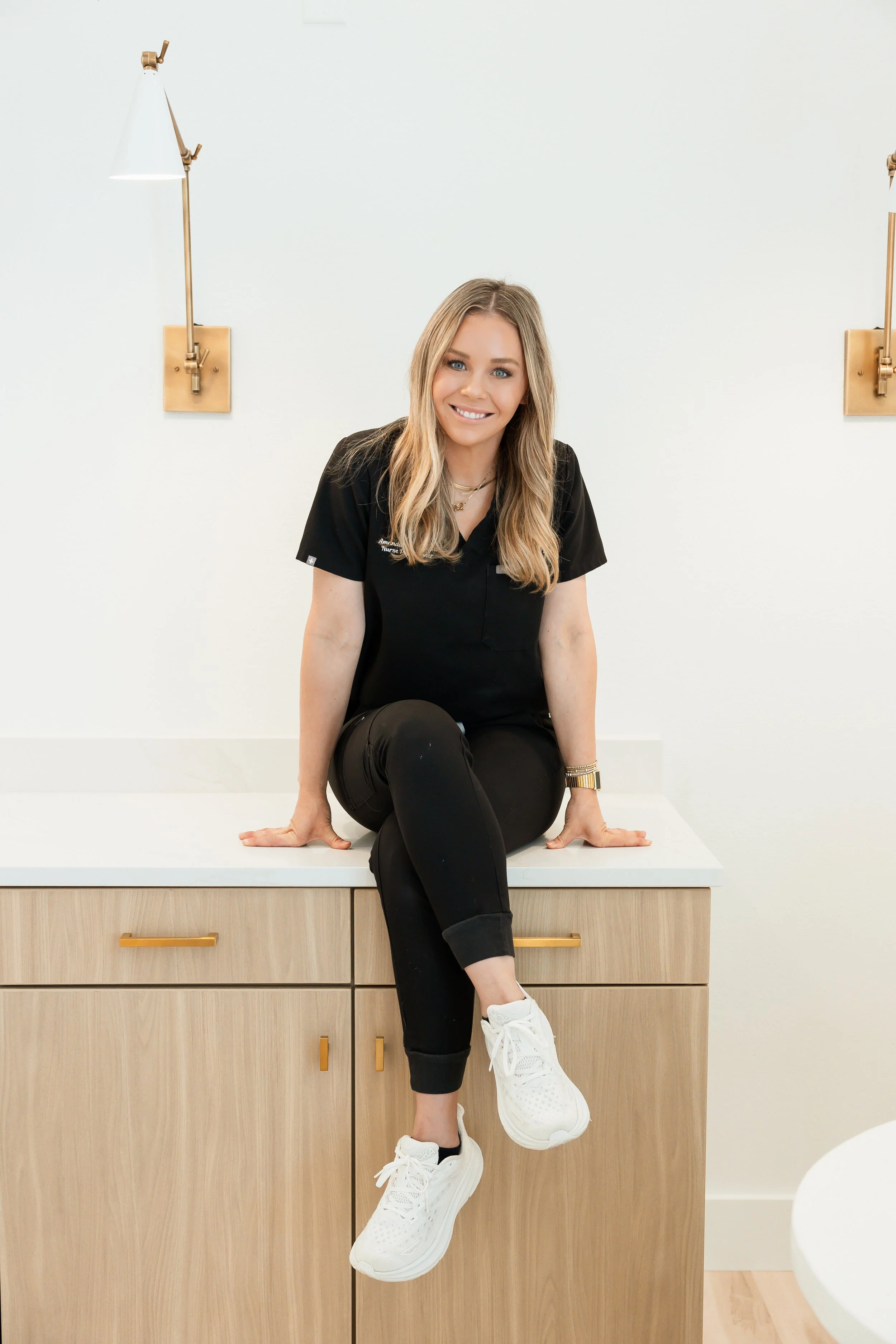Amanda Boatwright, owner and nurse practitioner at West Em Medical Spa, smiling while sitting on a white countertop wearing scrubs