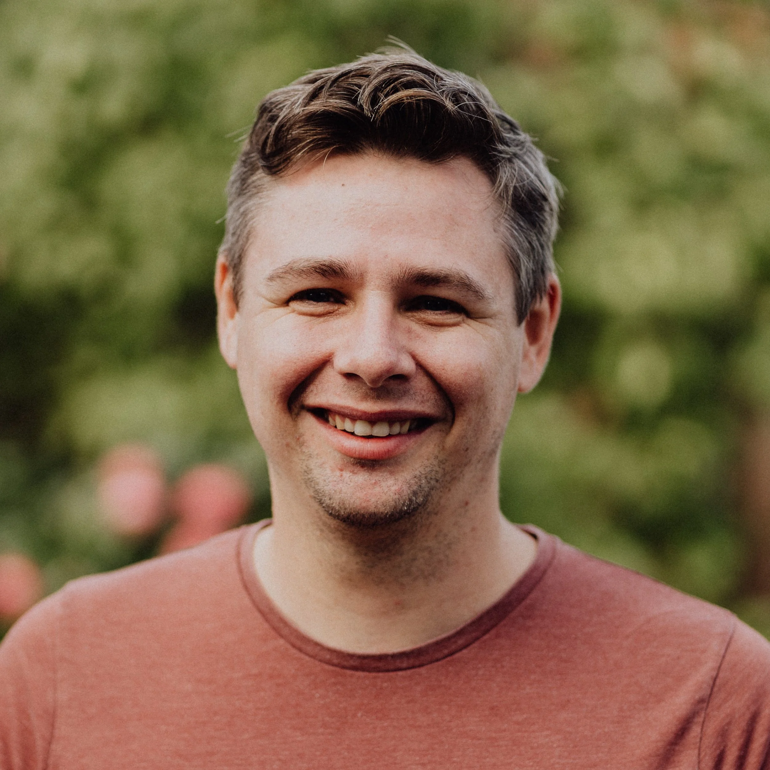 Lächelnder Mann im Freien mit grüner, verschwommener Natur im Hintergrund.