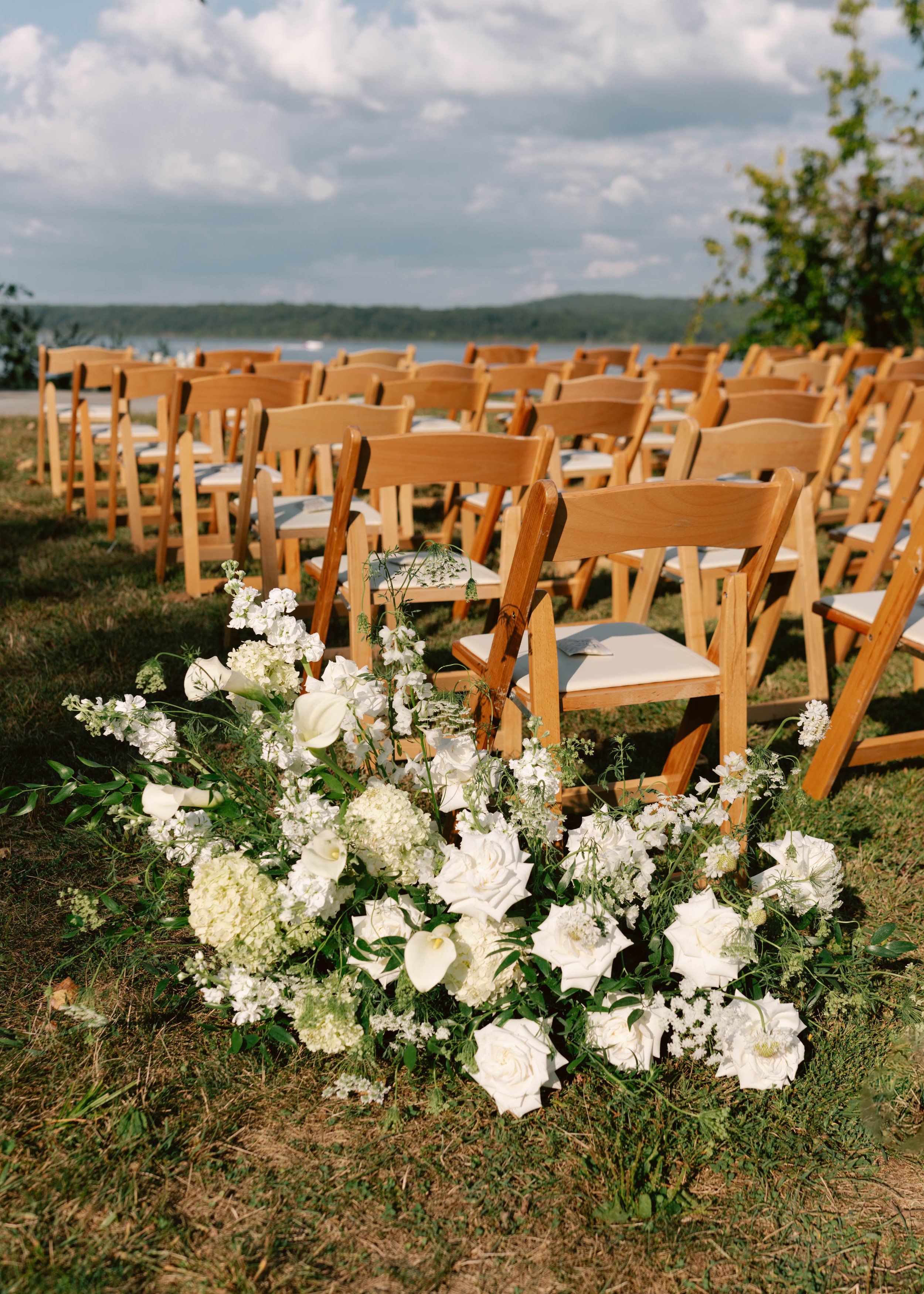 Wooden chairs arranged outdoors with a floral arrangement of white flowers in the foreground, set up for a wedding ceremony near a body of water under a partly cloudy sky.
