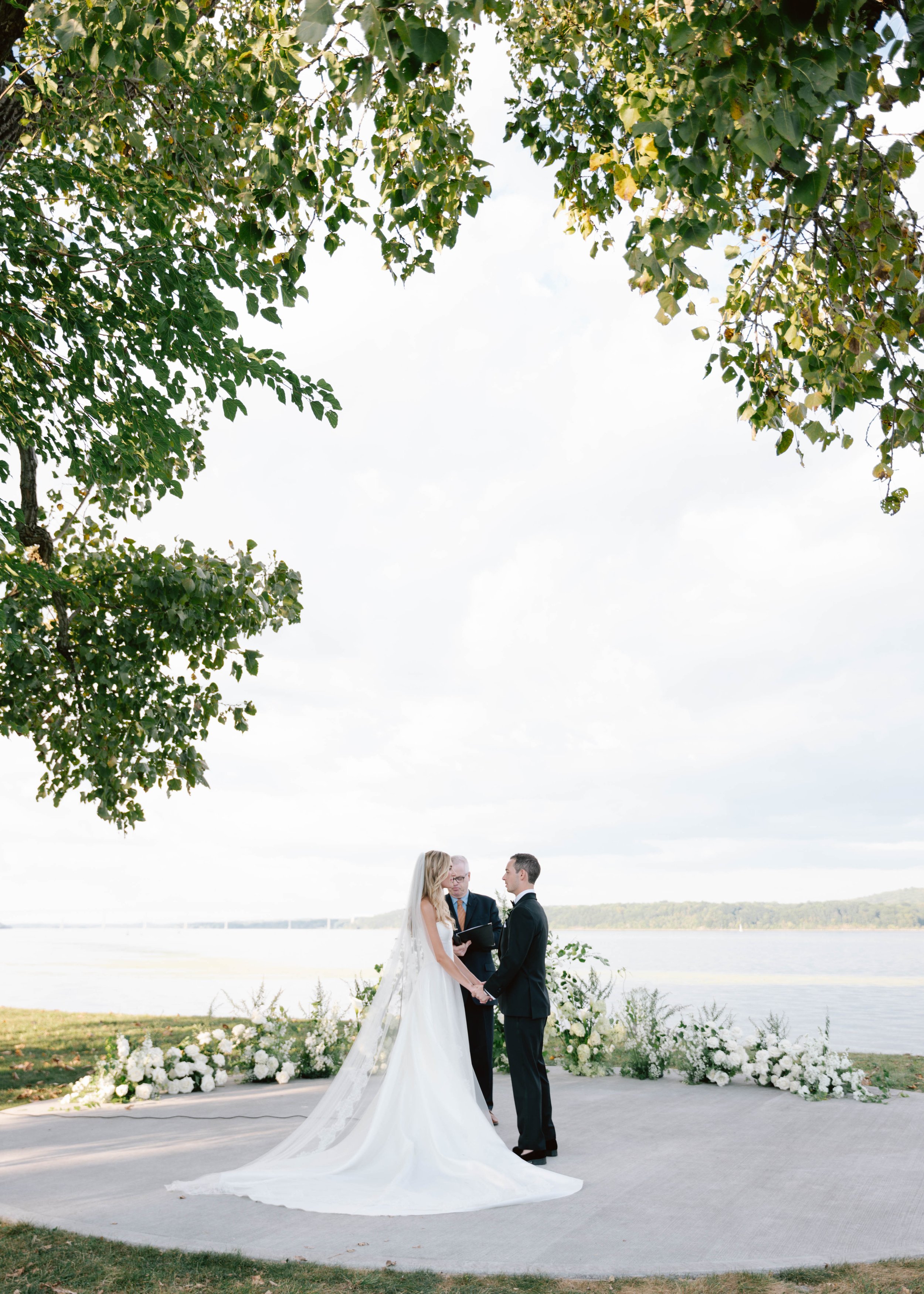A bride and groom holding hands during their outdoor wedding ceremony by a lake, officiant standing between them with floral arrangements in the background, under a large leafy tree.