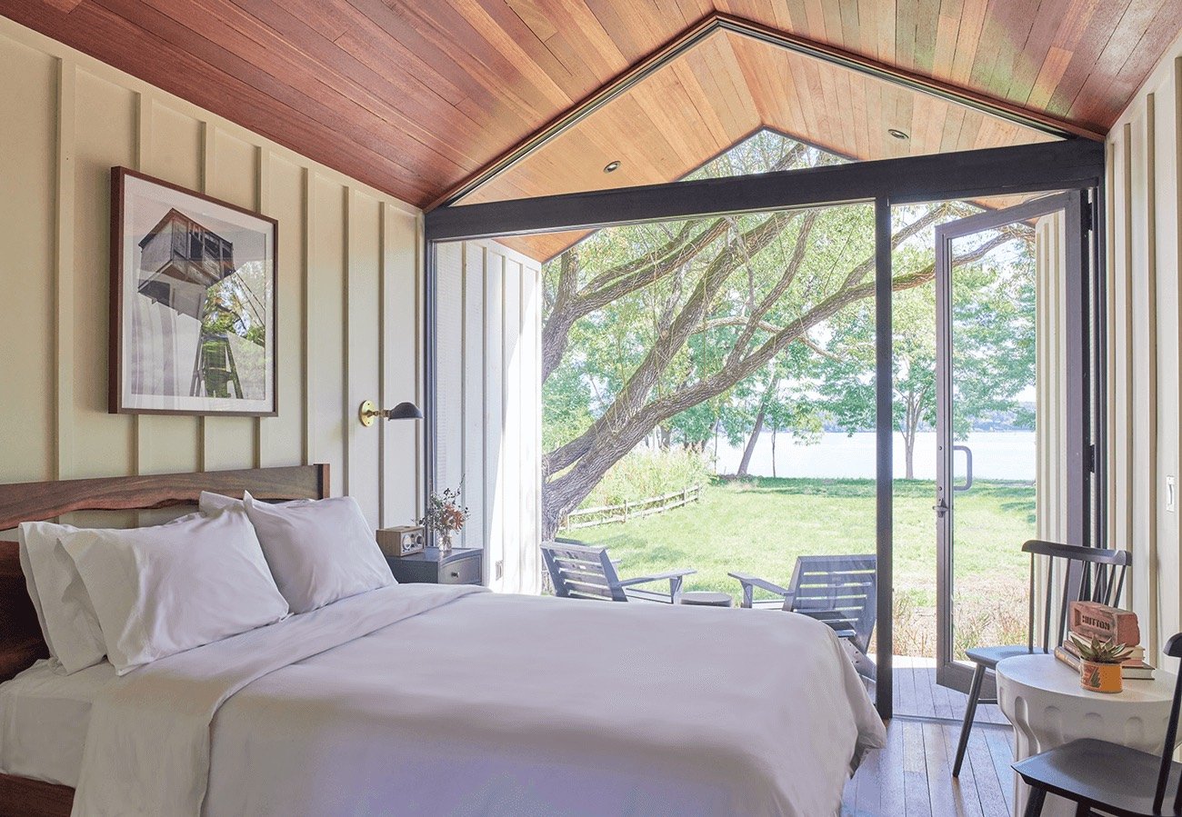 Bedroom with a large sliding glass door opening to a view of green grass, trees, and a body of water outside.