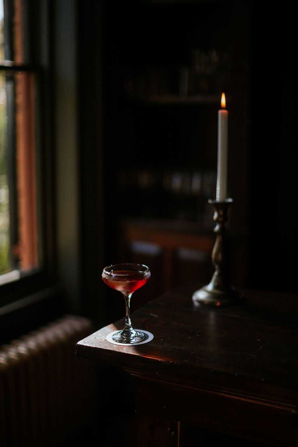 A glass of red wine on a wooden table next to a lit candle in a dark room, with a window showing trees outside.