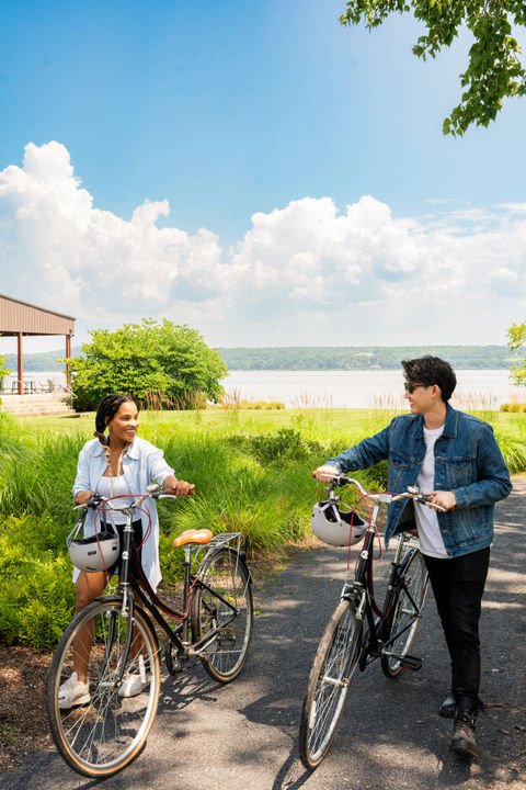 A man and woman with bicycles talking in a park near a lake under a blue sky with clouds.