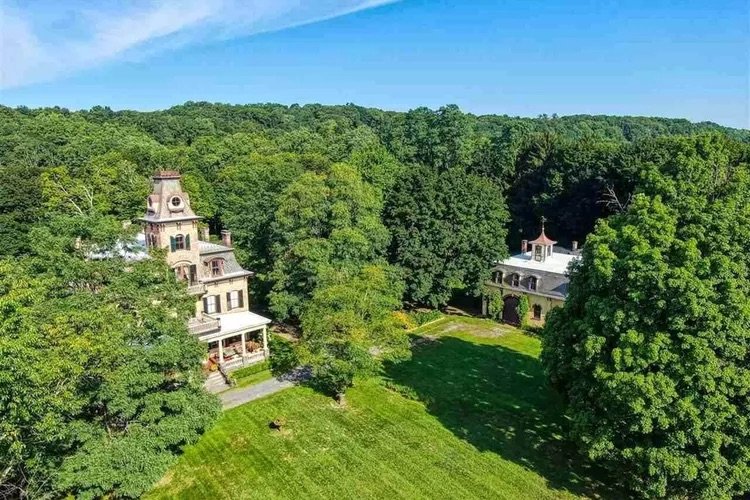 An aerial view of a large, Victorian-style house and a smaller building, surrounded by trees and grass in a wooded area on a clear day.