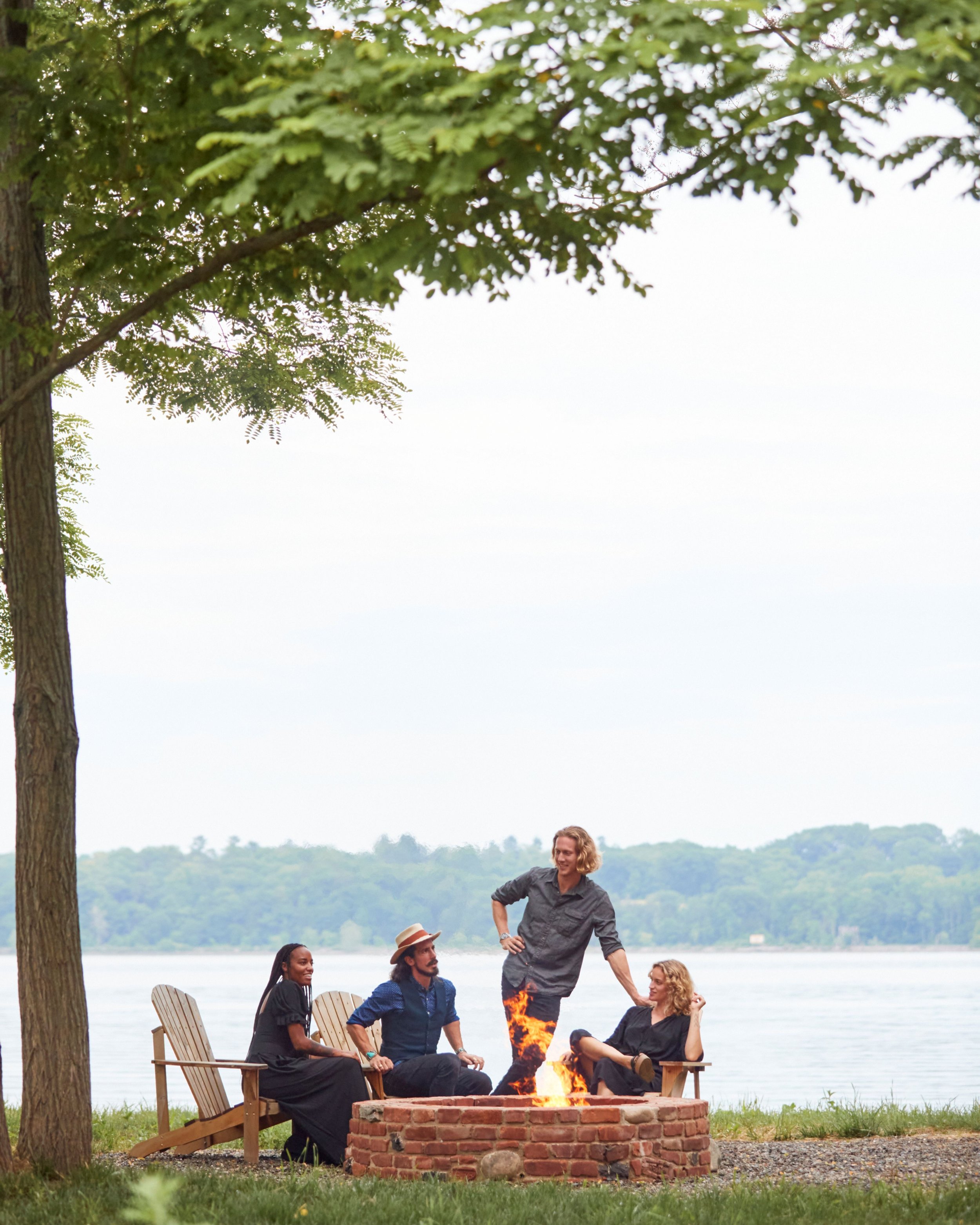 Four friends sitting and standing around a fire pit by a river, with trees and water in the background, enjoying a casual outdoor gathering.