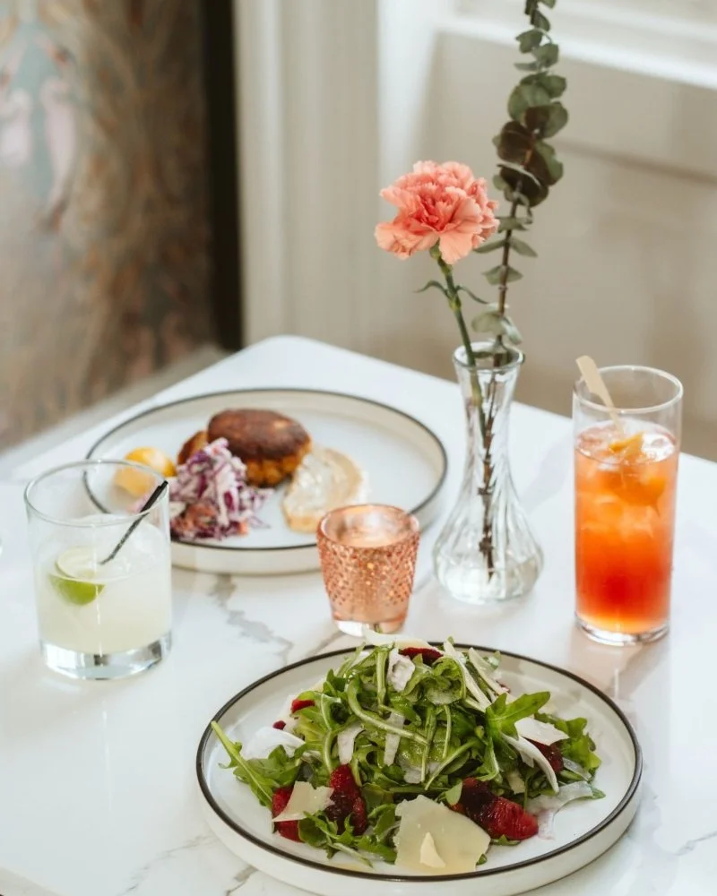 A chef wearing a black and white striped apron is placing a piece of dark red fruit or vegetable onto a dish with a spoon. The dish appears to be a dessert with fruits and sauce, garnished with seeds or spices, on a wooden countertop next to a plastic container of similar dark red items.