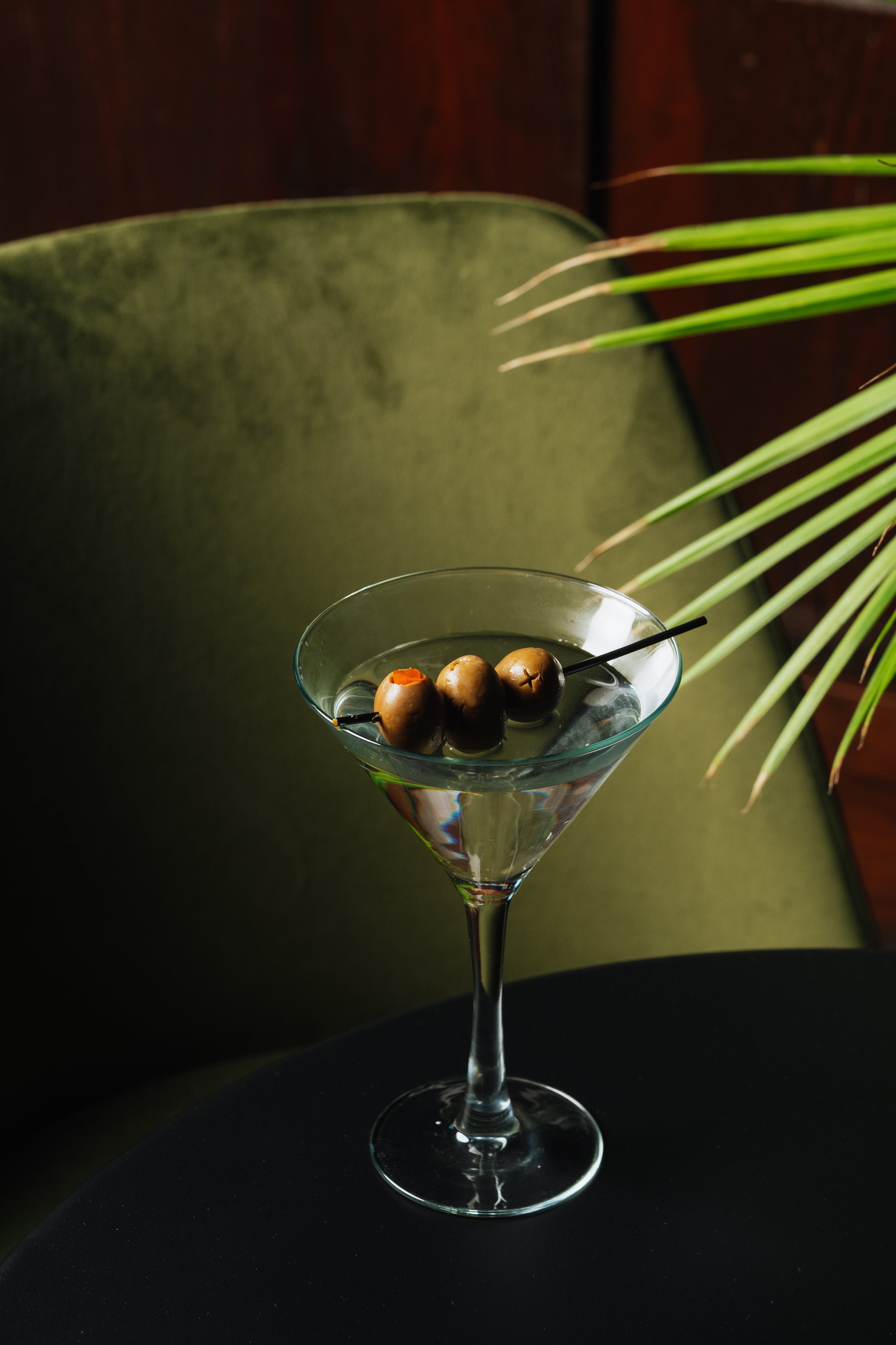 A martini glass filled with a clear liquid, garnished with three green olives on a skewer, placed on a black surface. A green upholstered chair and sunlight with a palm leaf are in the background.
