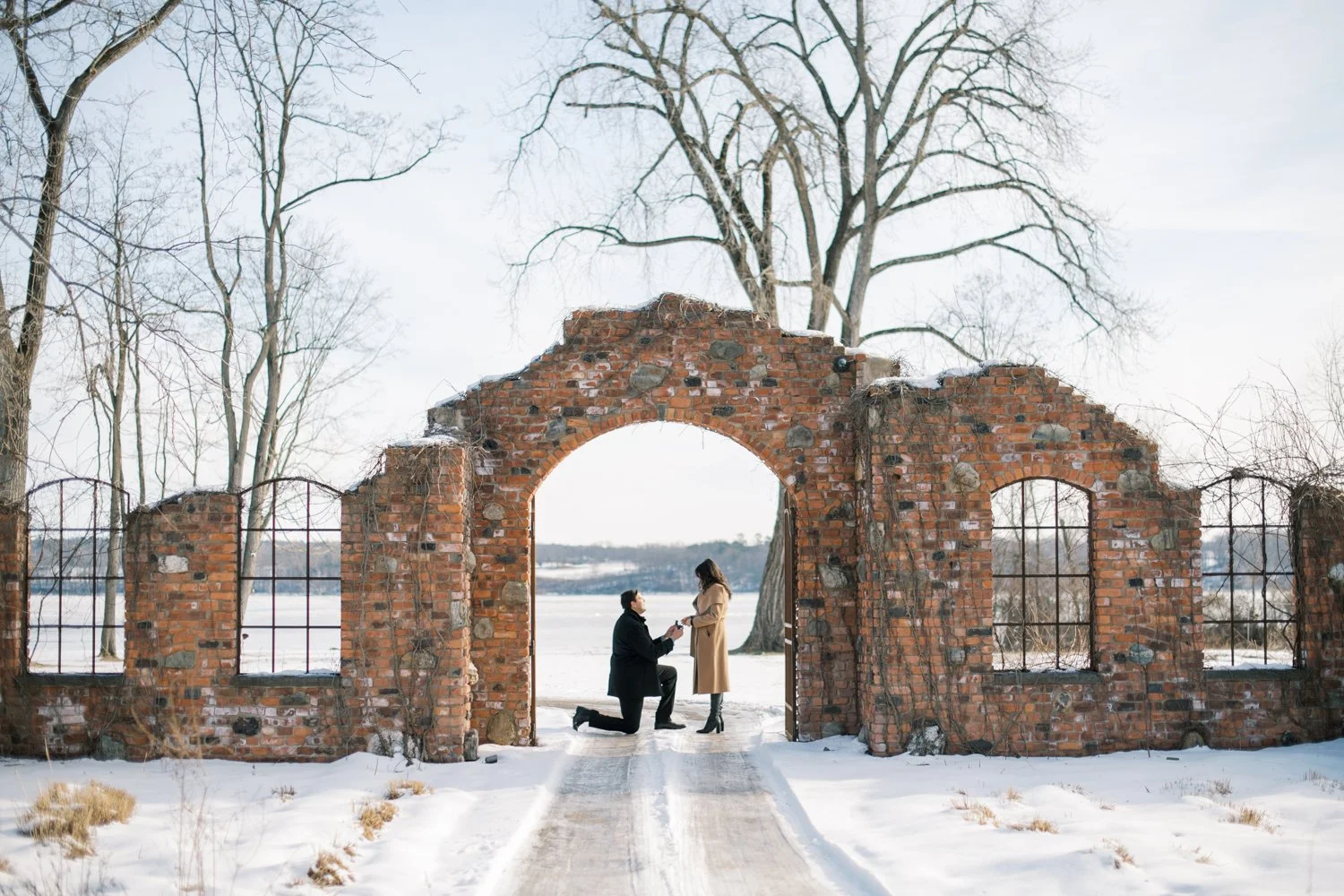 A happy couple wearing matching orange coats, holding hands, with the woman showing her engagement ring. They are outdoors in a park during fall.