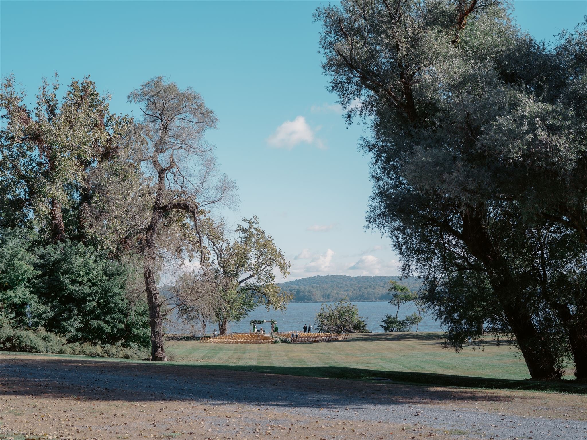 A scenic view of a park with large trees, a grassy area, a lake, and a small wedding ceremony setup with chairs and an arch in the distance under a blue sky.