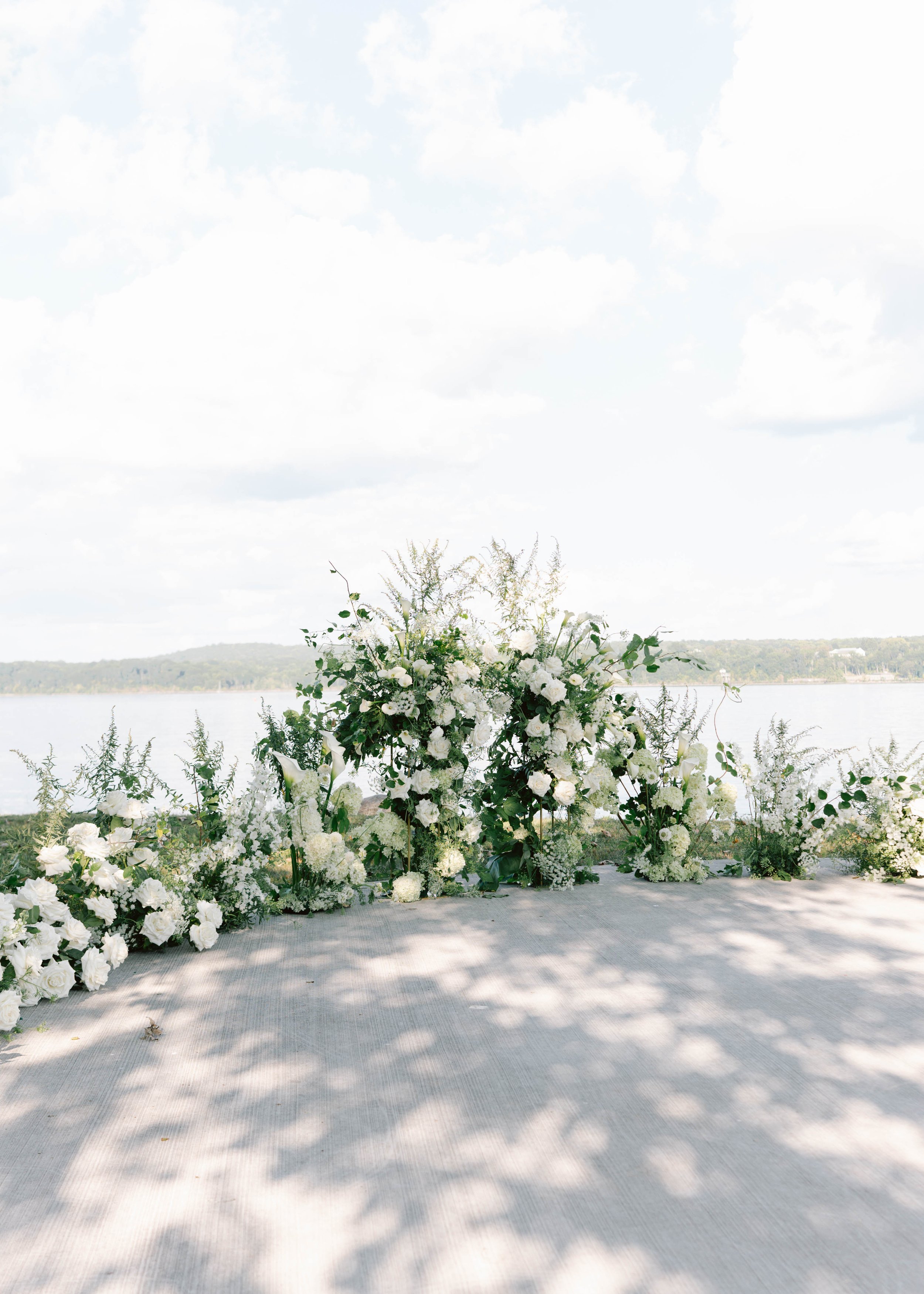 A floral wedding arch decorated with white flowers and greenery near a body of water with trees in the background.