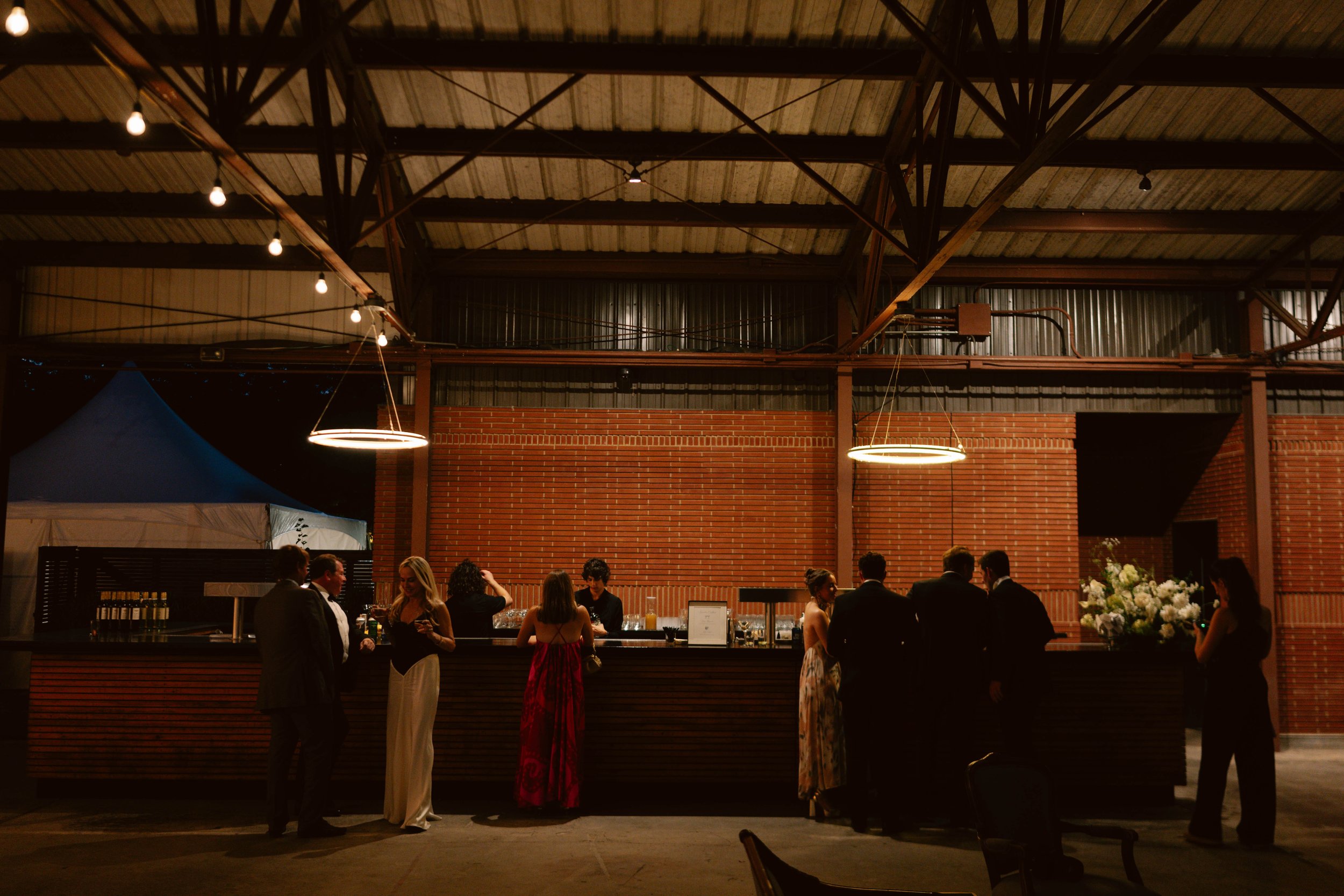 People at a bar in an industrial-style venue with brick walls and modern circular hanging lights.