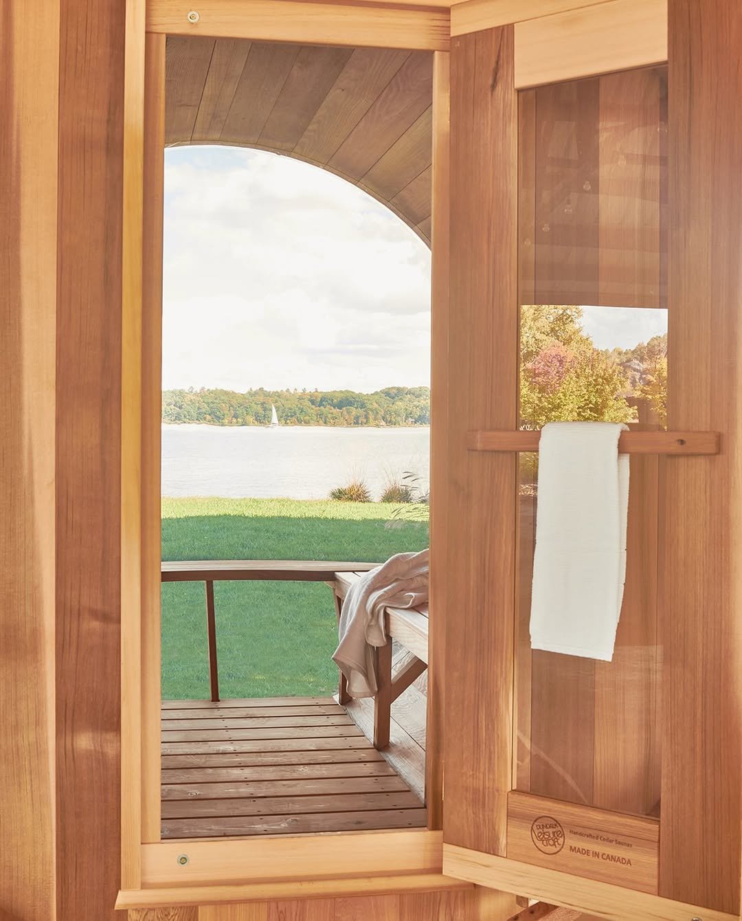 View through the open door of a wooden sauna cabin, showing a small wooden deck and a scenic view of the Hudson River with trees and a sailboat in the water.