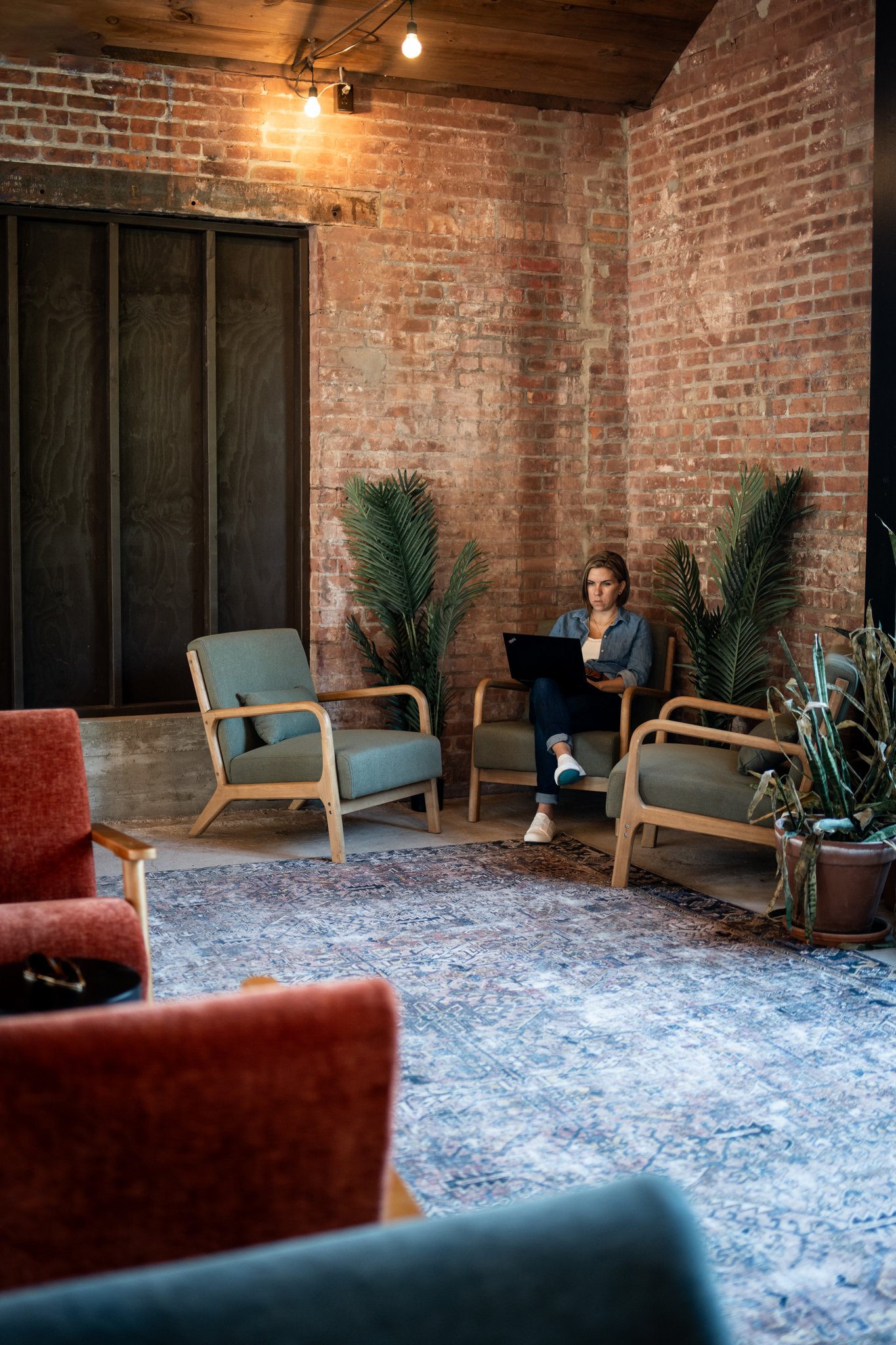 A woman sitting on a wooden armchair, working on a laptop in a cozy, industrial-style lounge with brick walls, several potted plants, and colorful chairs around a patterned rug.