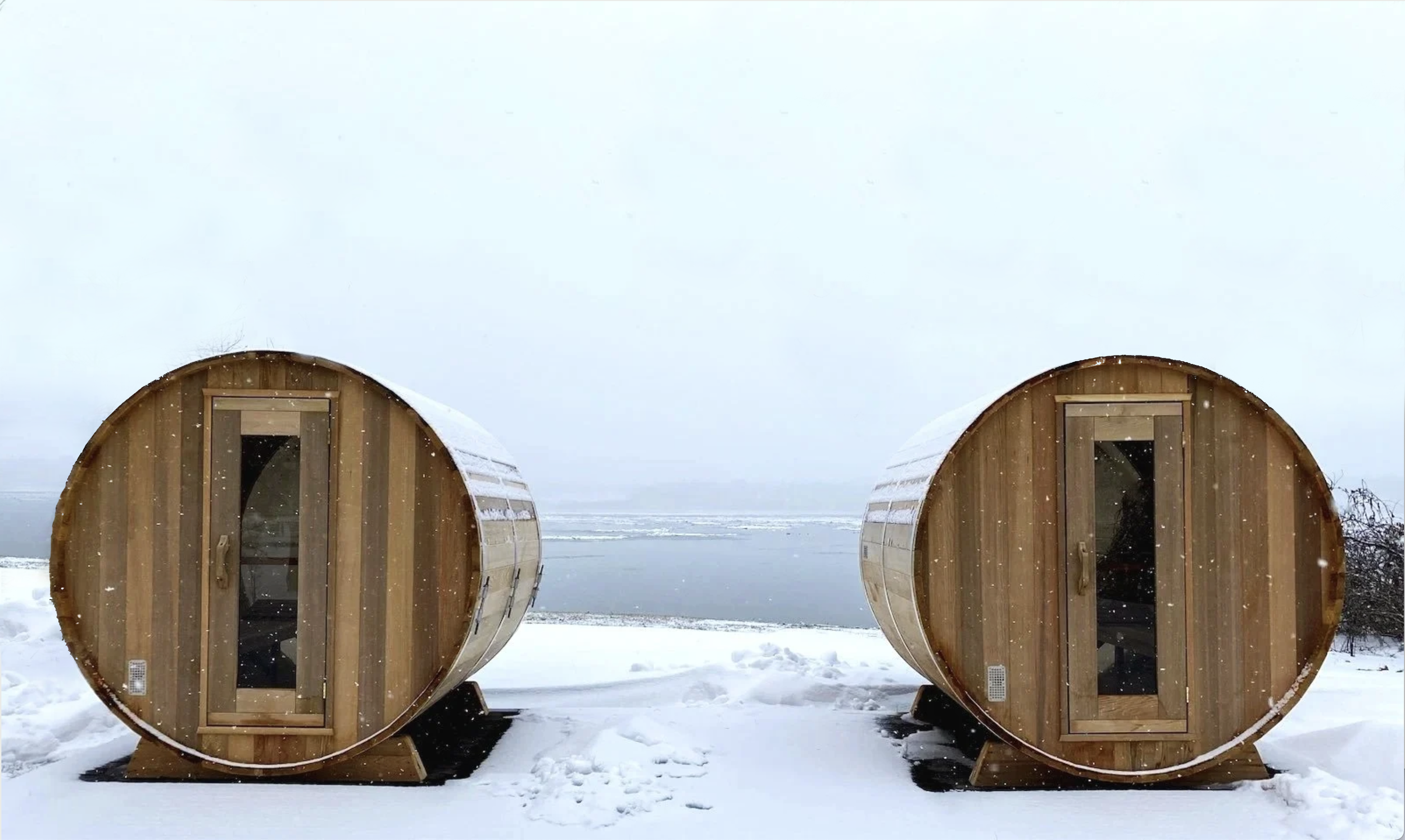 Two wooden barrel-shaped saunas on snow-covered ground with a body of water and cloudy sky in the background.