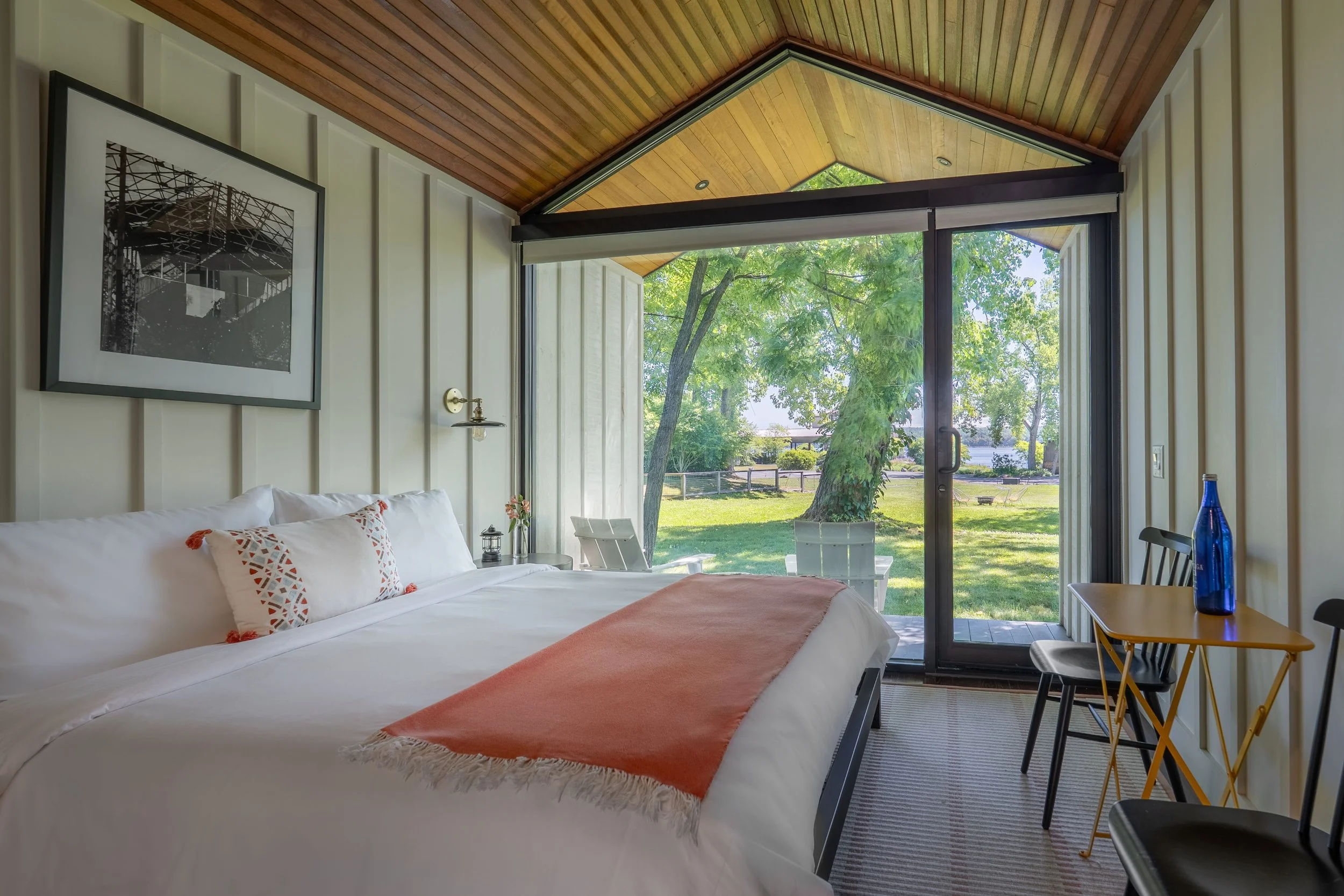 A bedroom with a large glass sliding door opening to a green outdoor yard with trees, a fence, and a patio. The room has a wooden ceiling, white paneled walls, a bed with white linens and orange accents, a bedside lamp, a small table with flowers, an