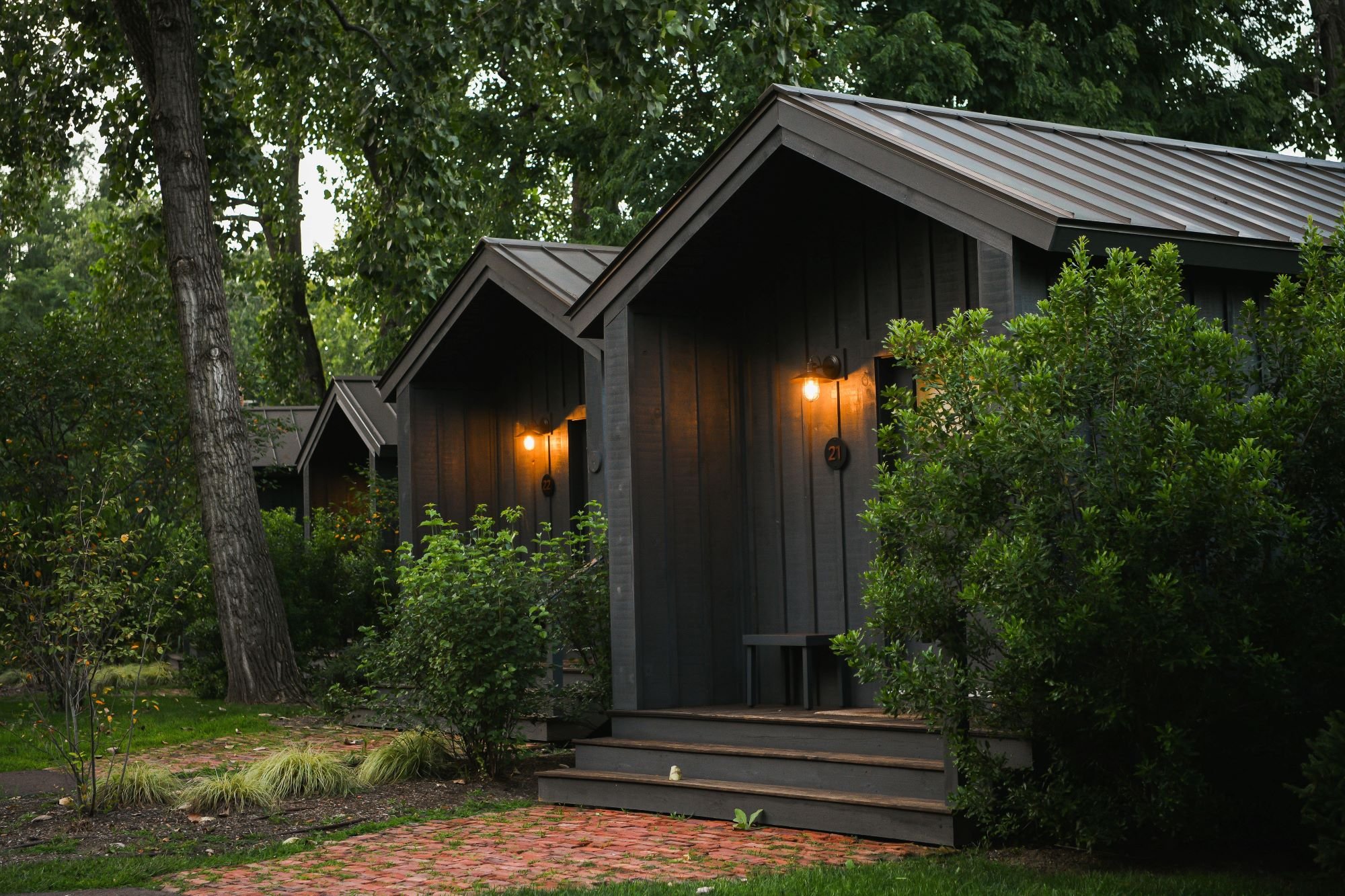 Three black wooden cottages with front steps, exterior lights on, surrounded by green trees and bushes, brick pathway leading to the entrance, in a wooded area.
