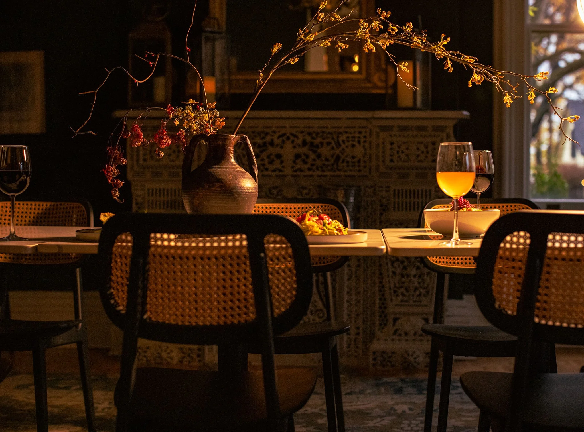 A dimly lit dining table with a decorative vase holding dried branches and flowers, two wine glasses with red and yellow drinks, and a bowl of food, with a window showing trees outside in the background. Aster restaurant, romantic dinner. 