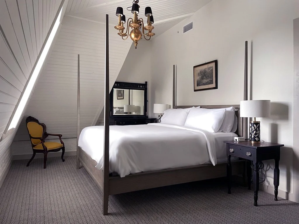 Interior of a bedroom with a four-poster bed, nightstand, yellow armchair, chandelier, and a framed picture on the wall.