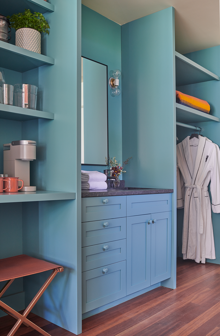 A modern bathroom nook with light blue cabinetry, a mirror, a light fixture with exposed light bulbs, and open shelves with towels and clothing, including a white robe, on a rod.