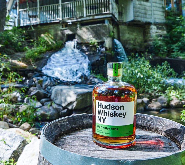 A bottle of Hudson Whiskey NY on a wooden barrel with a river and waterfall in the background.