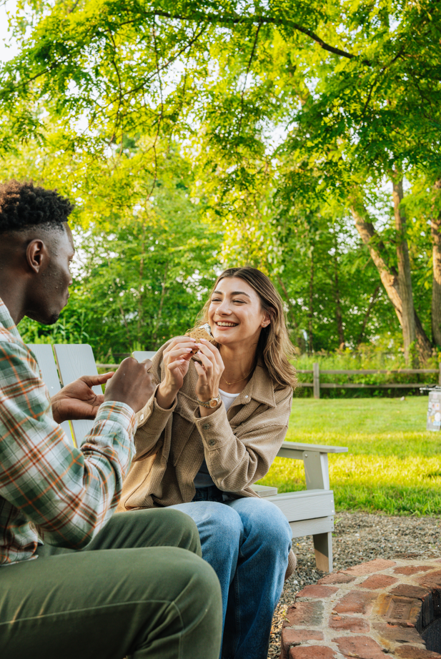 A man and woman sit on a park bench outdoors, sharing a snack surrounded by green trees and grass, enjoying a sunny day.