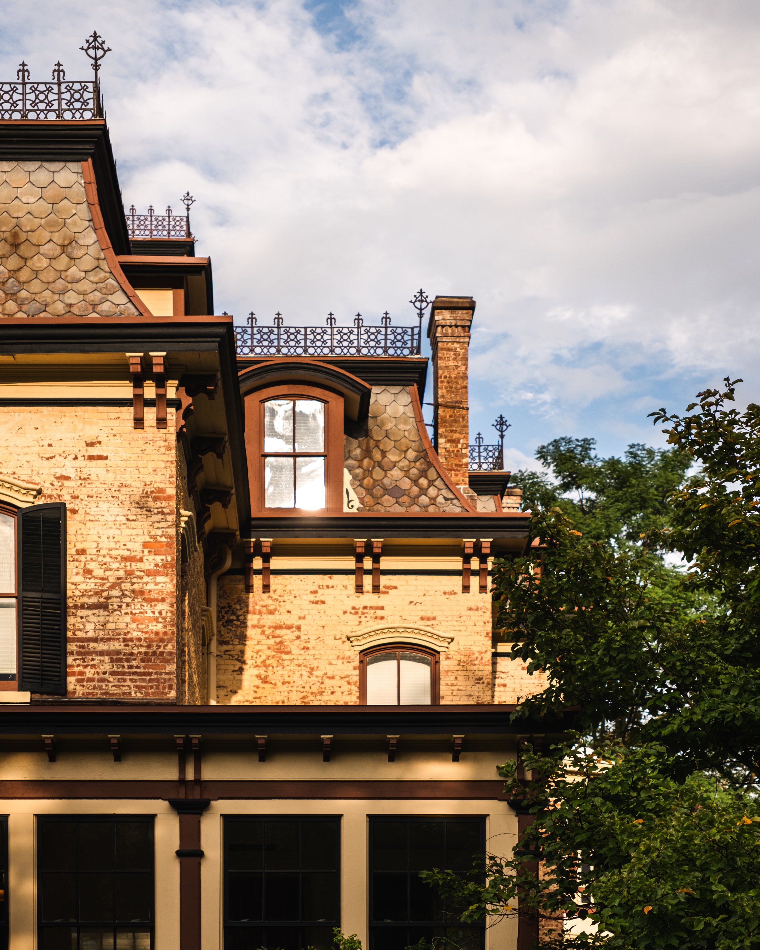 Close-up of a historic brick house with ornate ironwork on the roof and dormer windows under a partly cloudy sky, with trees nearby.