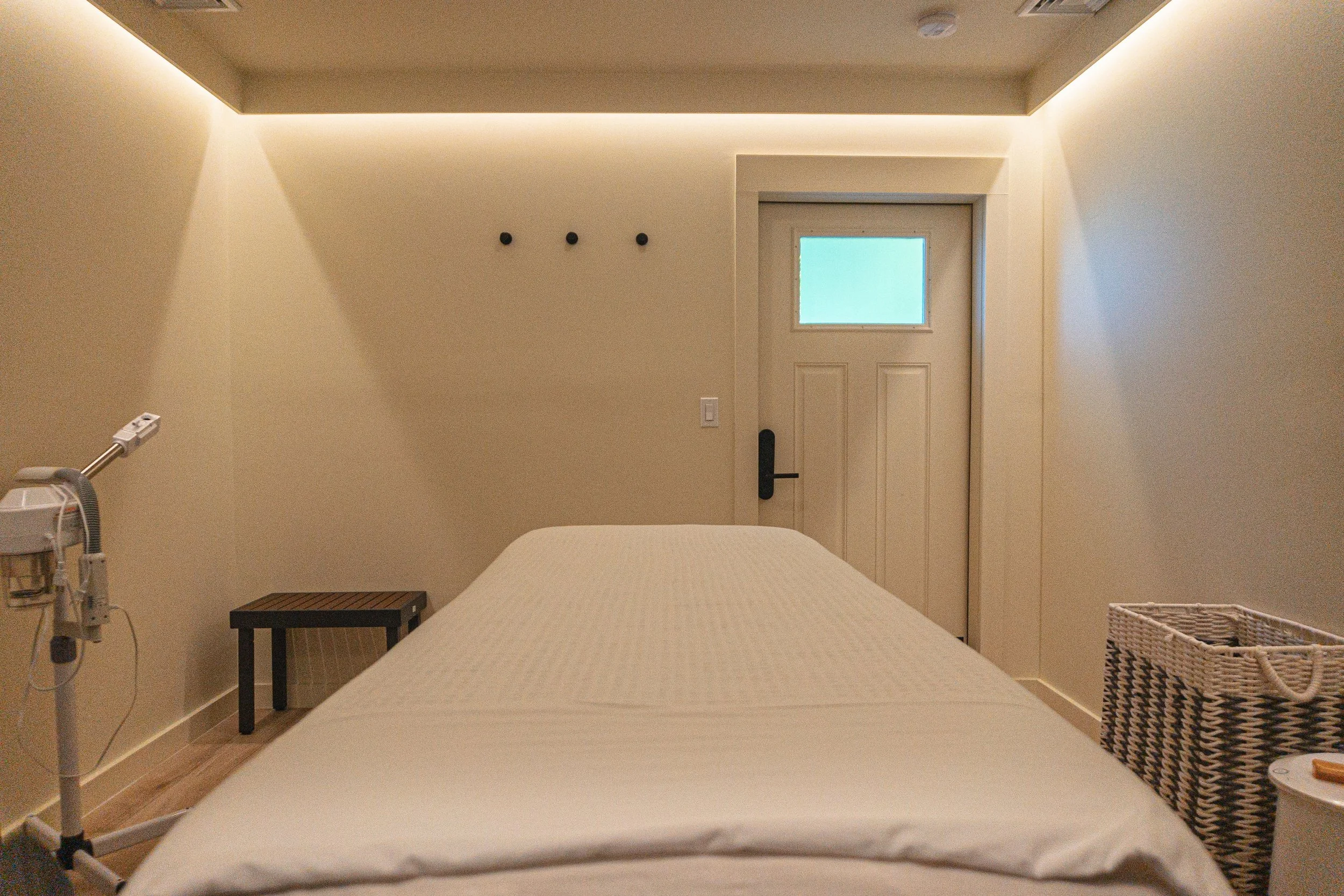 Treatment room with a bed, side table, wicker basket, and spa equipment, light-colored walls, and a door with a small window.